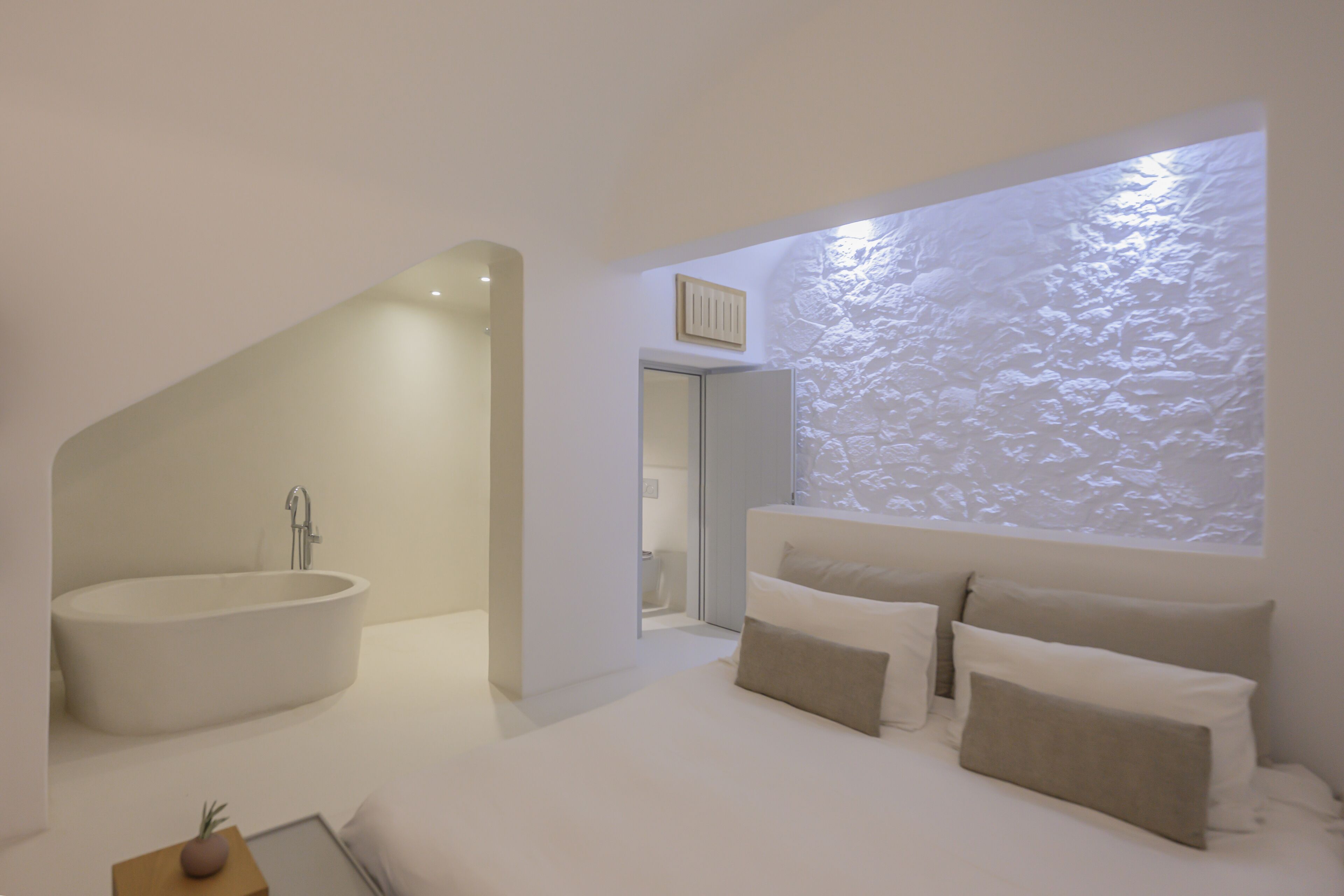 A modern bedroom with a large bathtub, a bed with multiple pillows, and a textured wall.