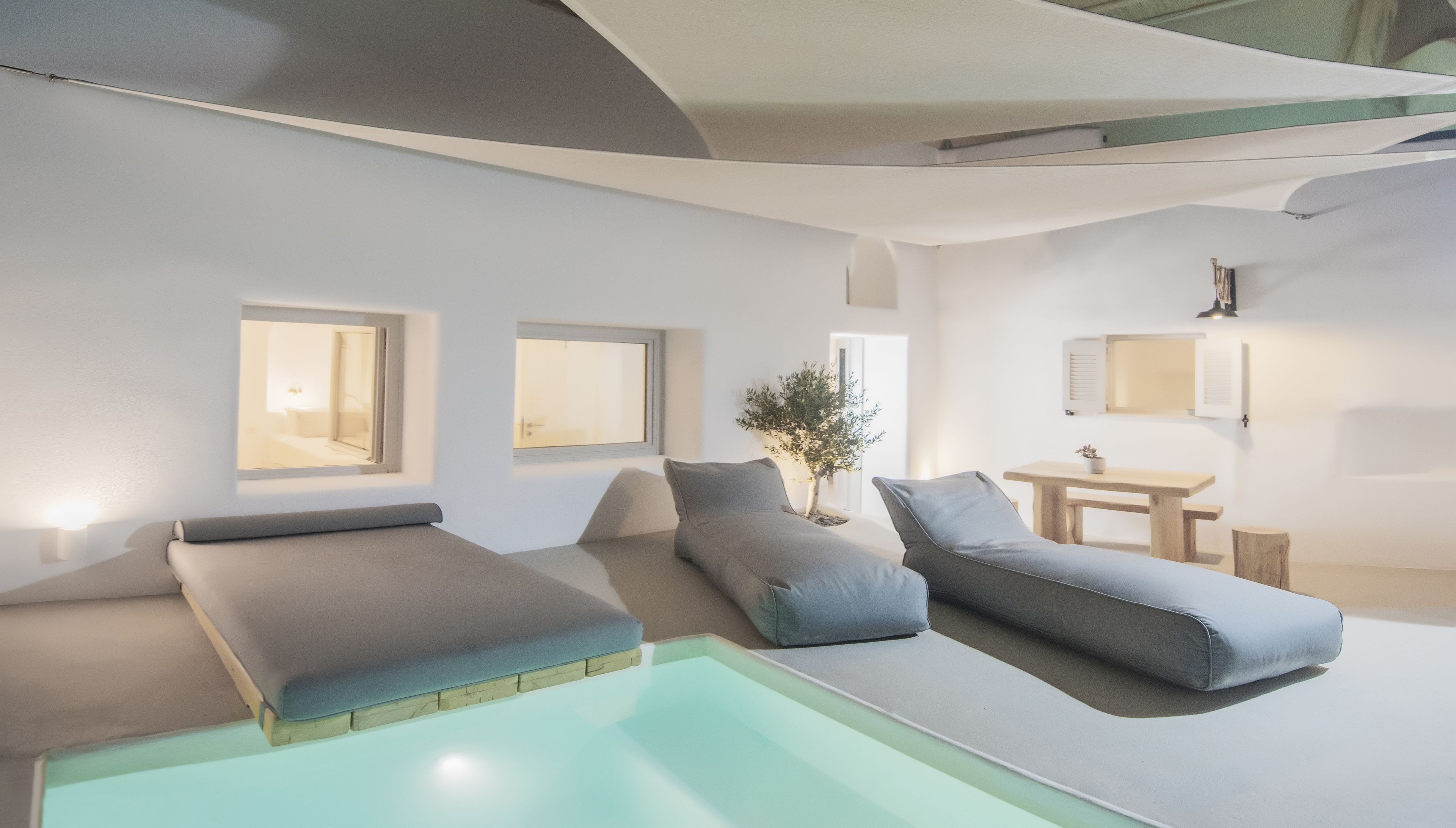 A modern spa room with a pool, two lounge chairs, a small table, and a potted plant.