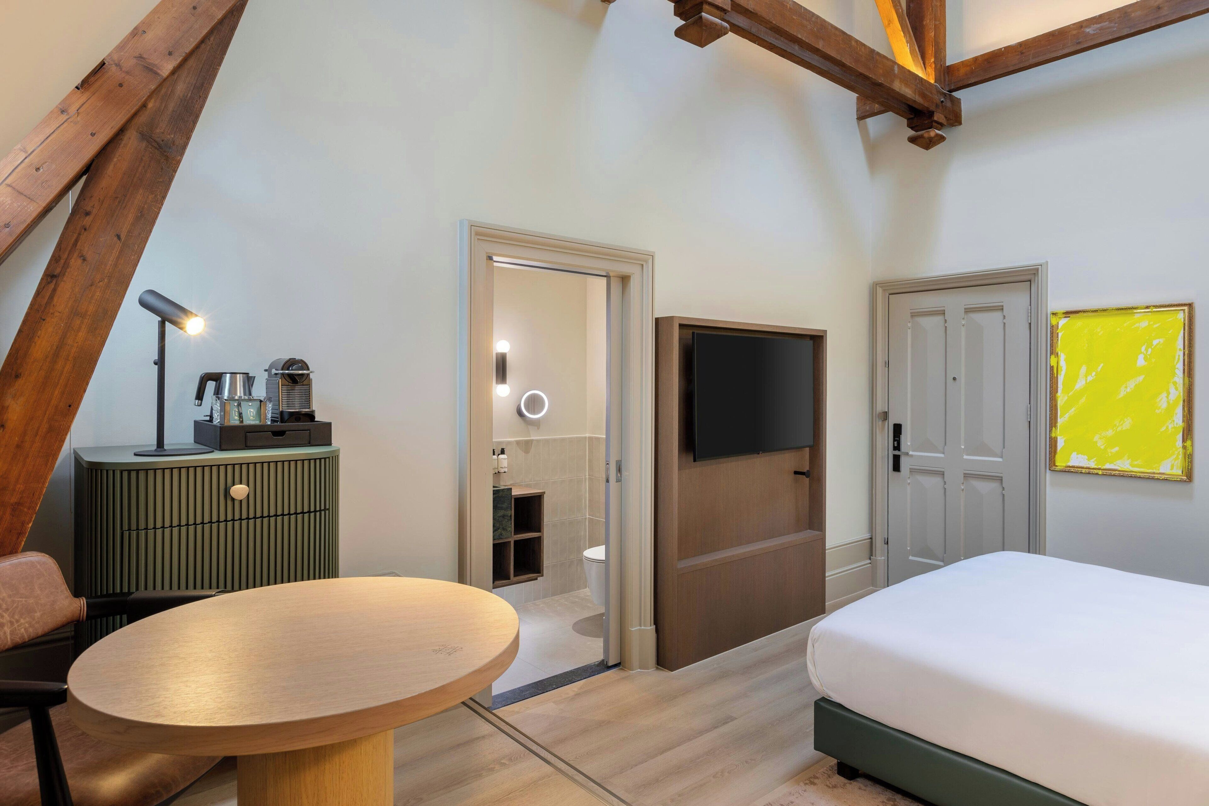 A modern hotel room with a wooden ceiling, a bed, a TV, a green minibar, and a round table.