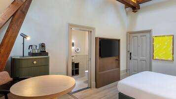A modern hotel room with a wooden ceiling, a bed, a TV, a green minibar, and a round table.