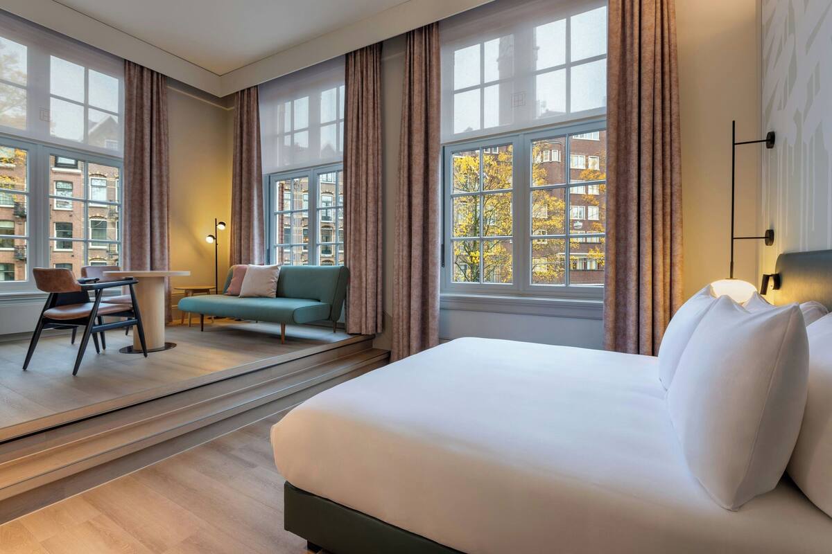 A hotel room with a large bed, a desk, a chair, and a sofa. There are large windows with curtains, and the room has a modern design.