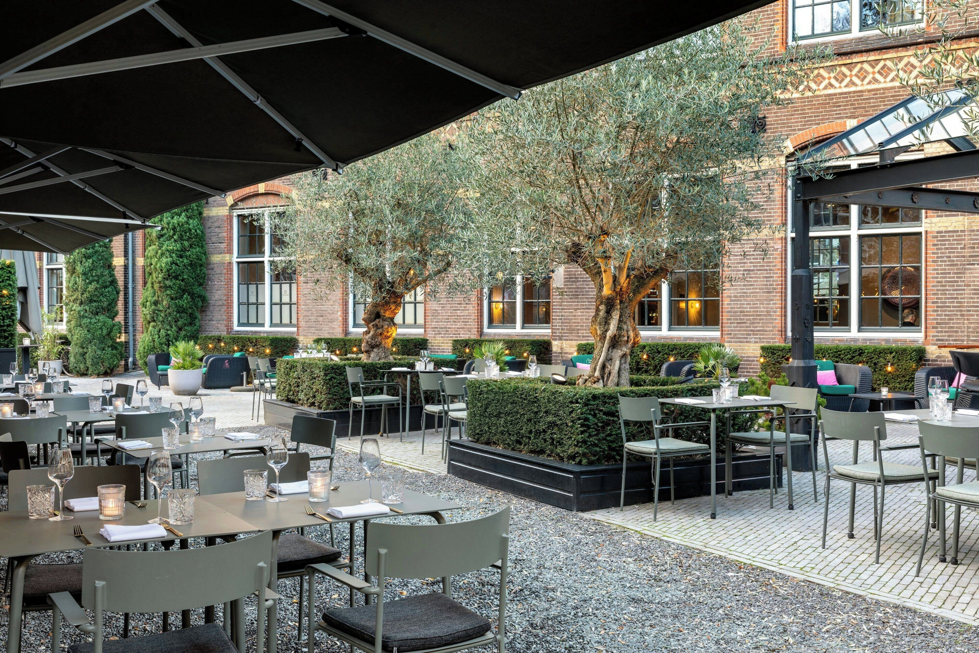 Een terras met tafels en stoelen, omgeven door groen en bakstenen muren.
