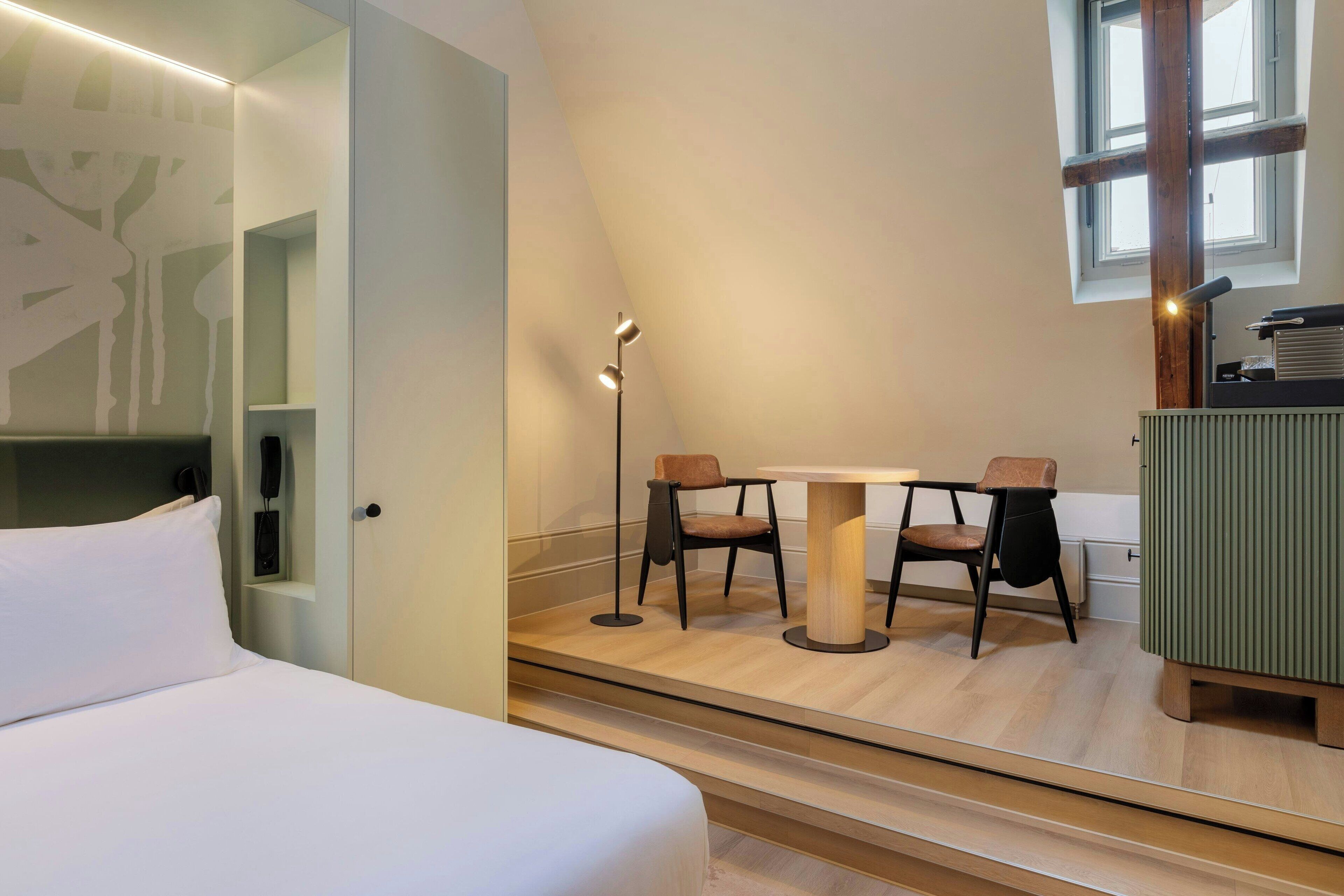 Una habitación de hotel con una cama, un escritorio con dos sillas, una lámpara de pie y un ventanal grande.