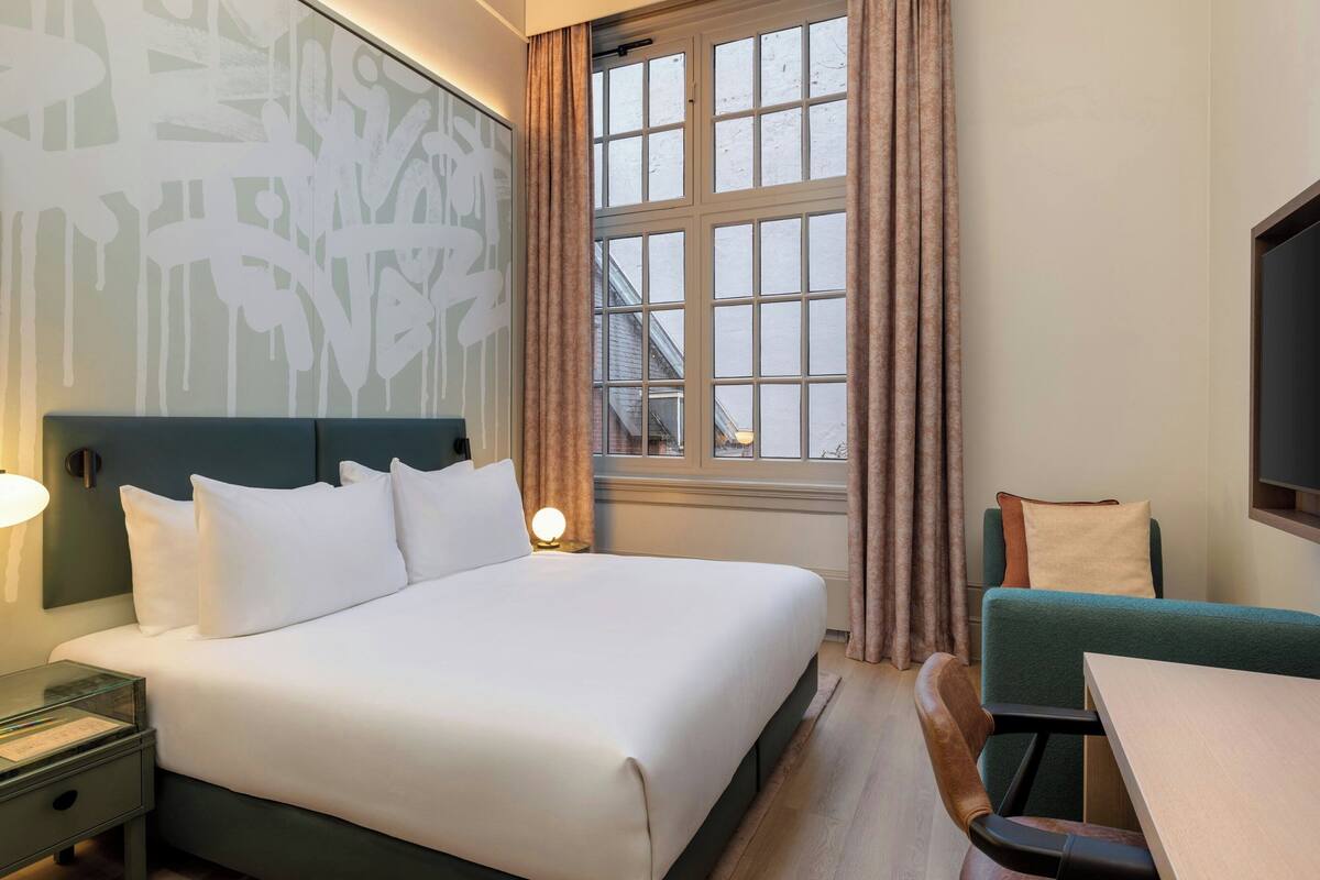 A hotel room with a large bed, a desk, a chair, and a window with curtains.