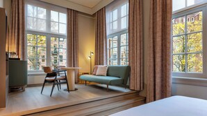 A hotel room with a bed, a green sofa, a chair, a small table, and a radiator. - The College Hotel Amsterdam, Autograph Collection (Amsterdam)