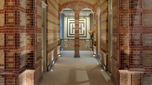 A corridor with brick walls and a central column, leading to a door with a geometric pattern. - The College Hotel Amsterdam, Autograph Collection (Amsterdam)