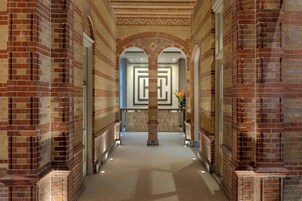 A corridor with brick walls and a central column, leading to a door with a geometric pattern.