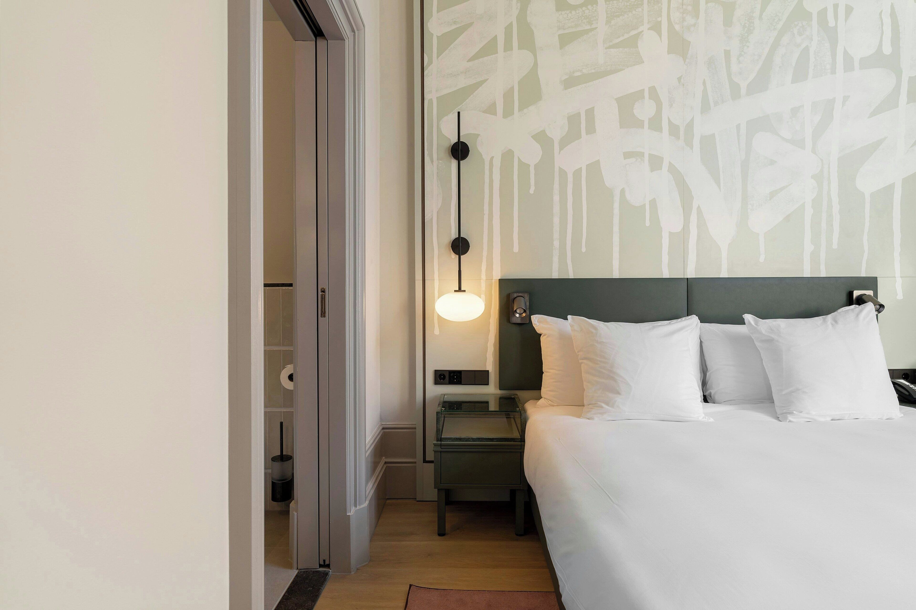 A hotel room with a bed, a nightstand, a wall with abstract art, and a hanging light fixture.