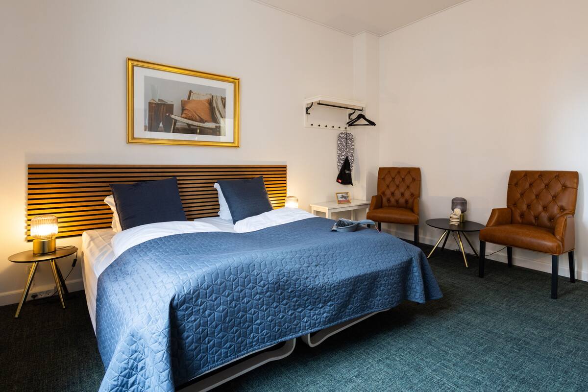 A hotel room with a bed, two chairs, a small table, a lamp, and a framed picture on the wall.