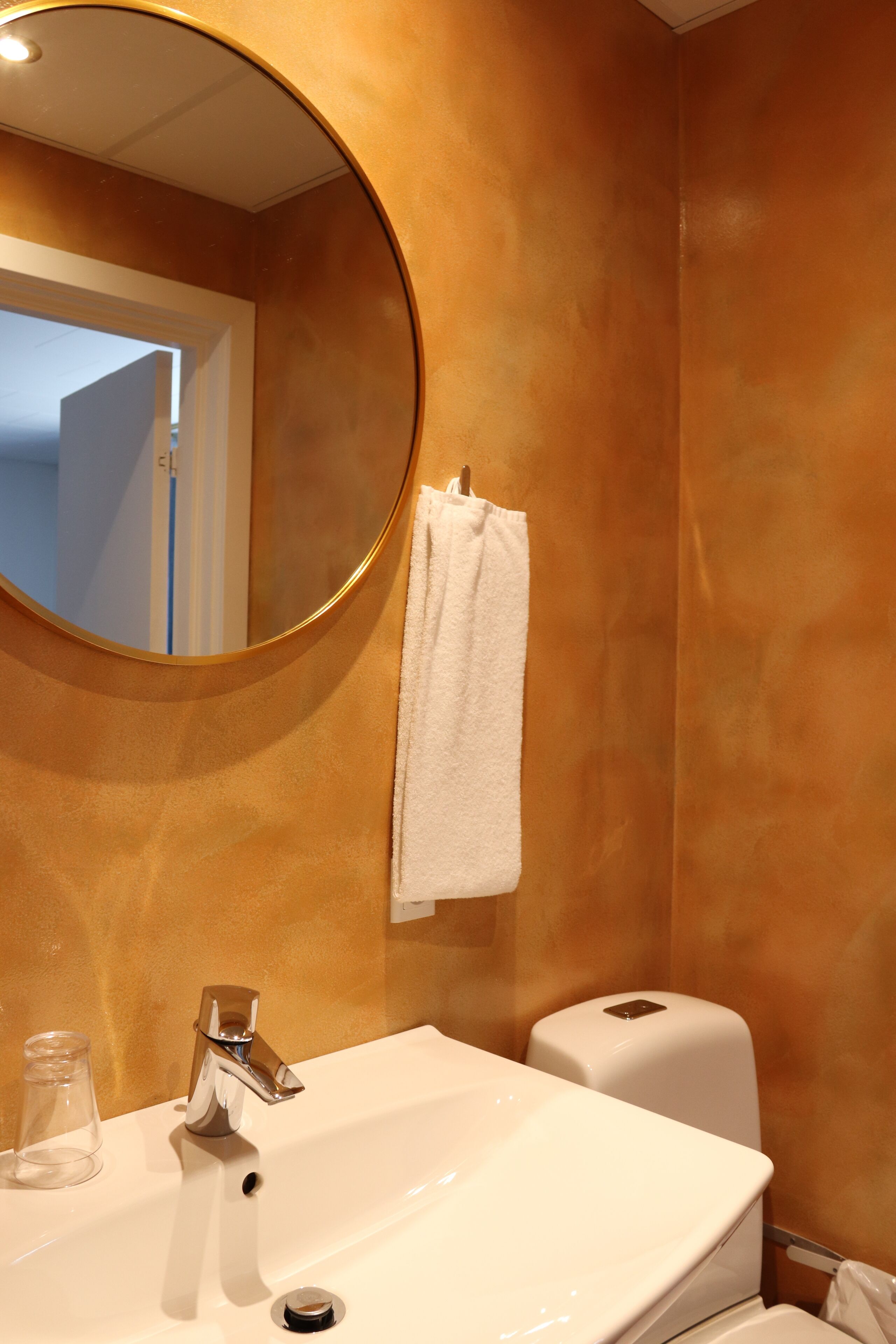 A bathroom with a round mirror, a white sink, a towel hanging on the wall, and a toilet paper dispenser.