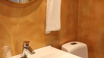 A bathroom with a round mirror, a white sink, a towel hanging on the wall, and a toilet paper dispenser.