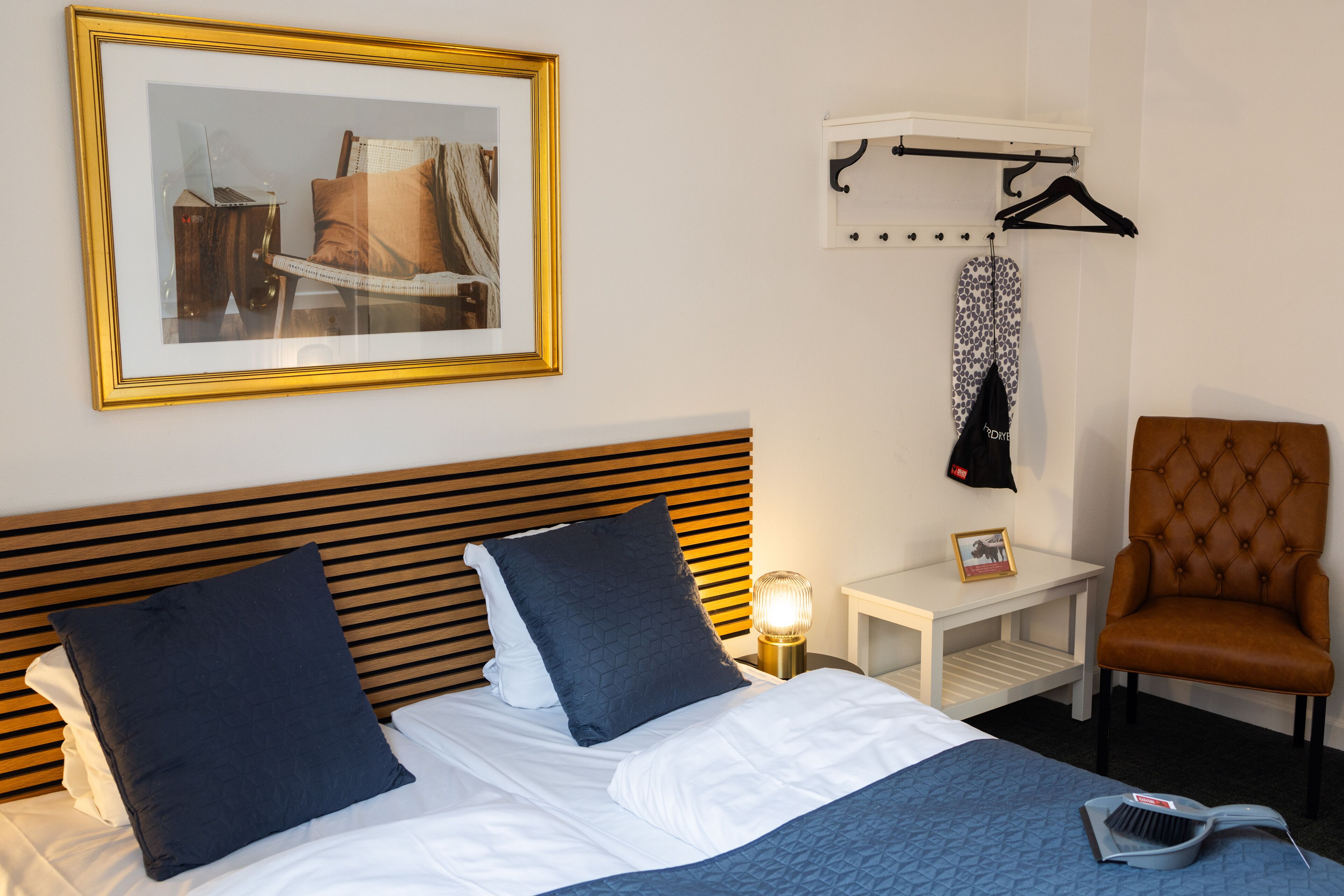 A neatly made bed with two pillows, a bedside table with a lamp, a chair, and a framed picture on the wall.