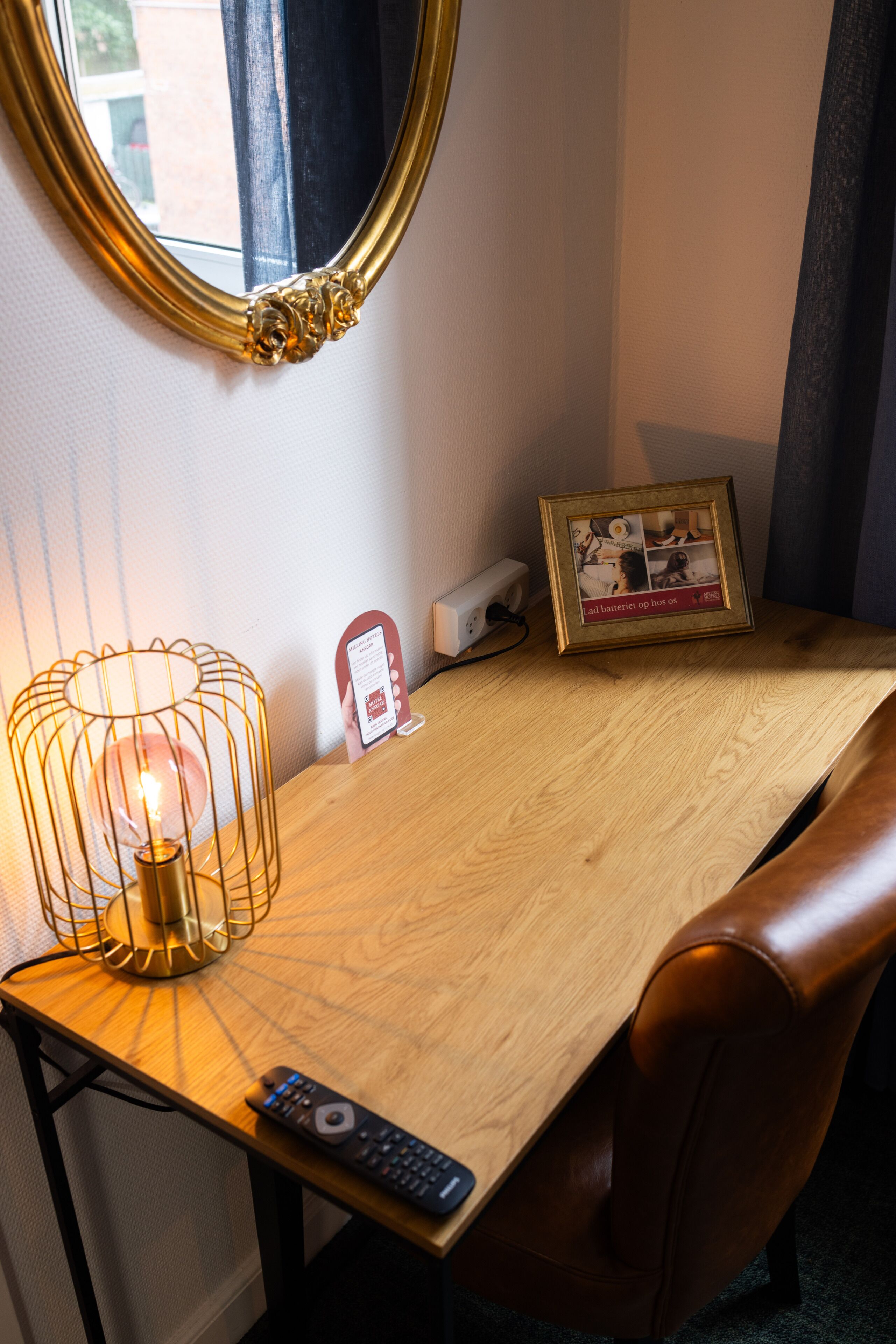 Een houten bureau met een lamp, een ingelijst beeld, een afstandsbediening en een stopcontact.