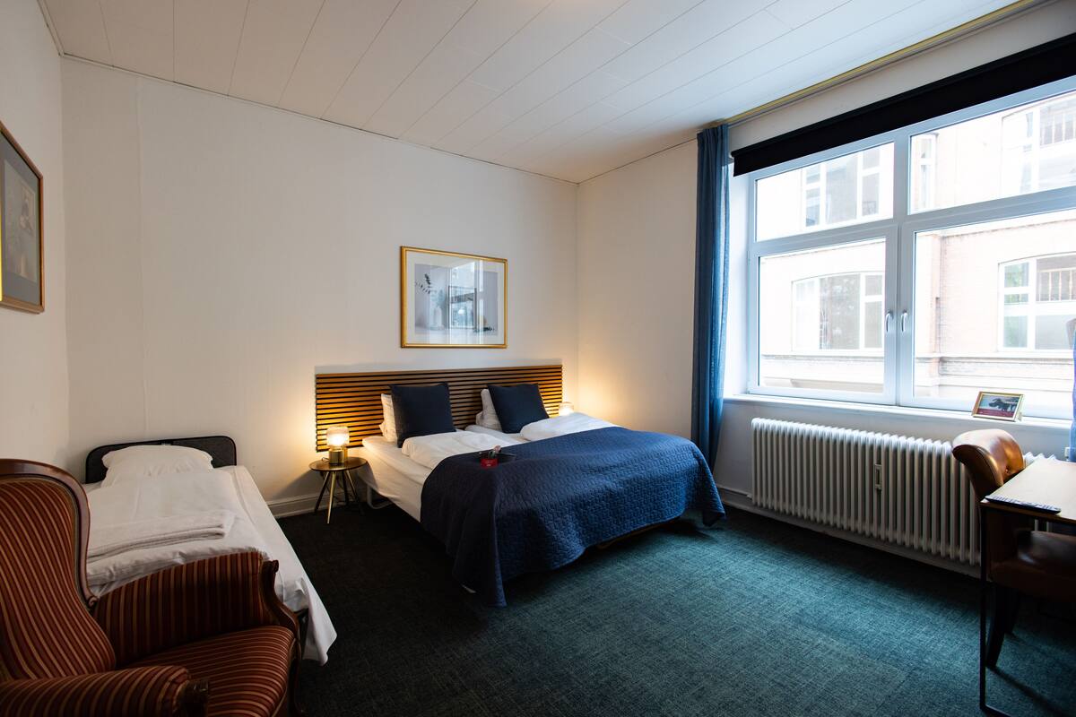 A hotel room with a bed, a chair, a desk, and a window with a view of buildings.