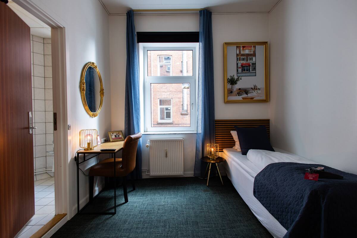A hotel room with a bed, a desk with a chair, a window with curtains, a radiator, and a framed picture on the wall.