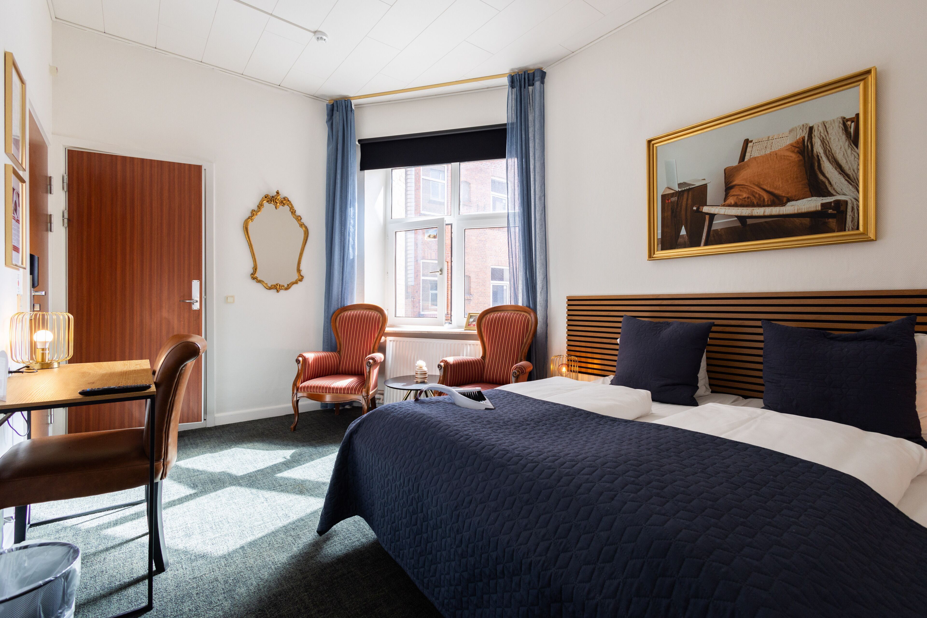 A hotel room with a large bed, a desk, two chairs, a window with curtains, and a framed picture on the wall.