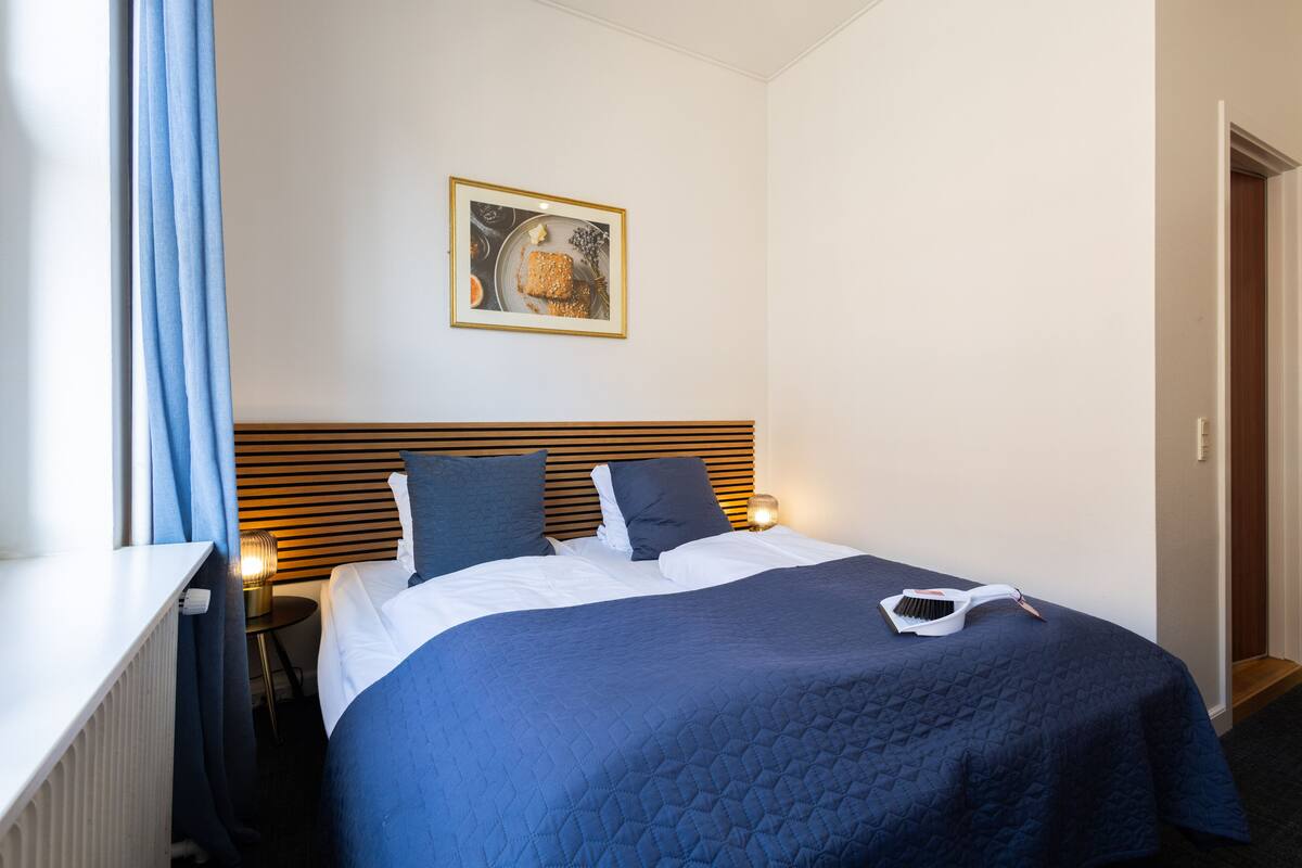 A neatly made bed with a blue quilt, two pillows, and a white blanket. There is a framed picture on the wall above the bed.