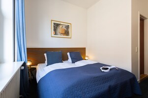 Una cama bien hecha con colcha azul, dos almohadas y una manta blanca. Encima de la cama, en la pared, hay un cuadro enmarcado.