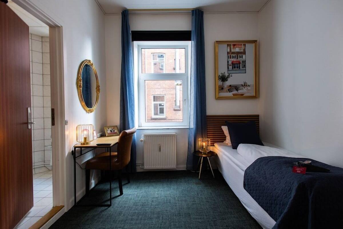 A hotel room with a bed, a desk, a chair, a radiator, a window with curtains, and a framed picture on the wall.