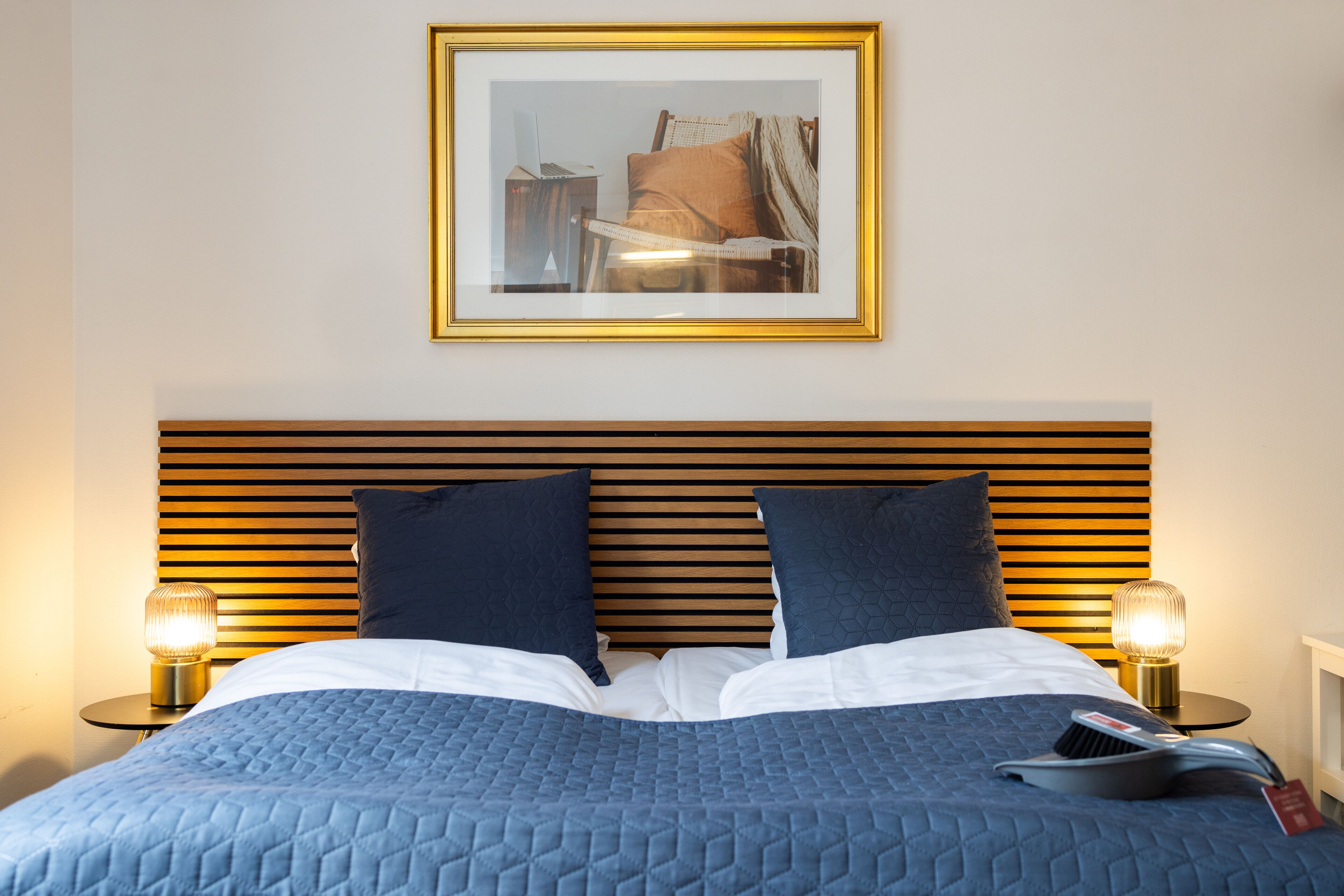 A neatly made bed with a blue quilt, two pillows, and a bedside table with a lamp.