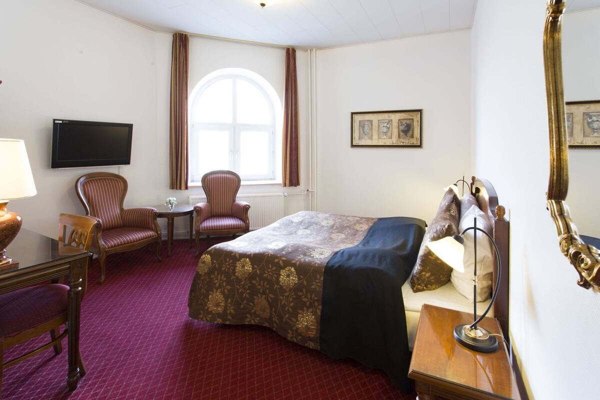 A hotel room with a bed, two chairs, a desk, a lamp, a television, and a window with curtains.
