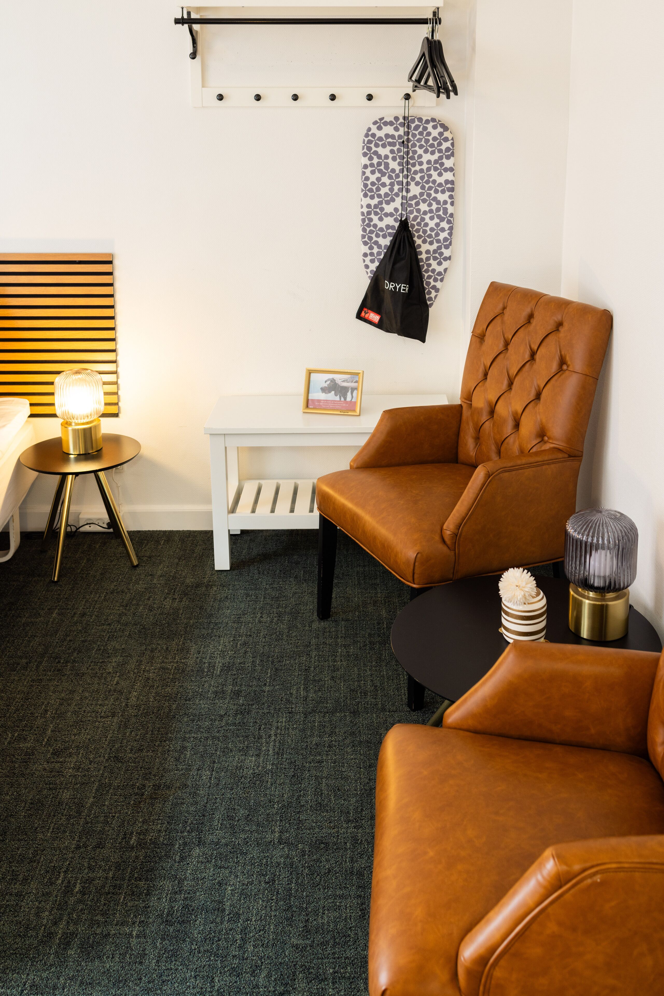 Ein Zimmer mit einem braunen Ledersessel, einem kleinen weißen Tisch mit Bilderrahmen, einem schwarzen Beistelltisch mit Dekorationsobjekten und einem Wandhaken mit einer gemusterten Tasche.
