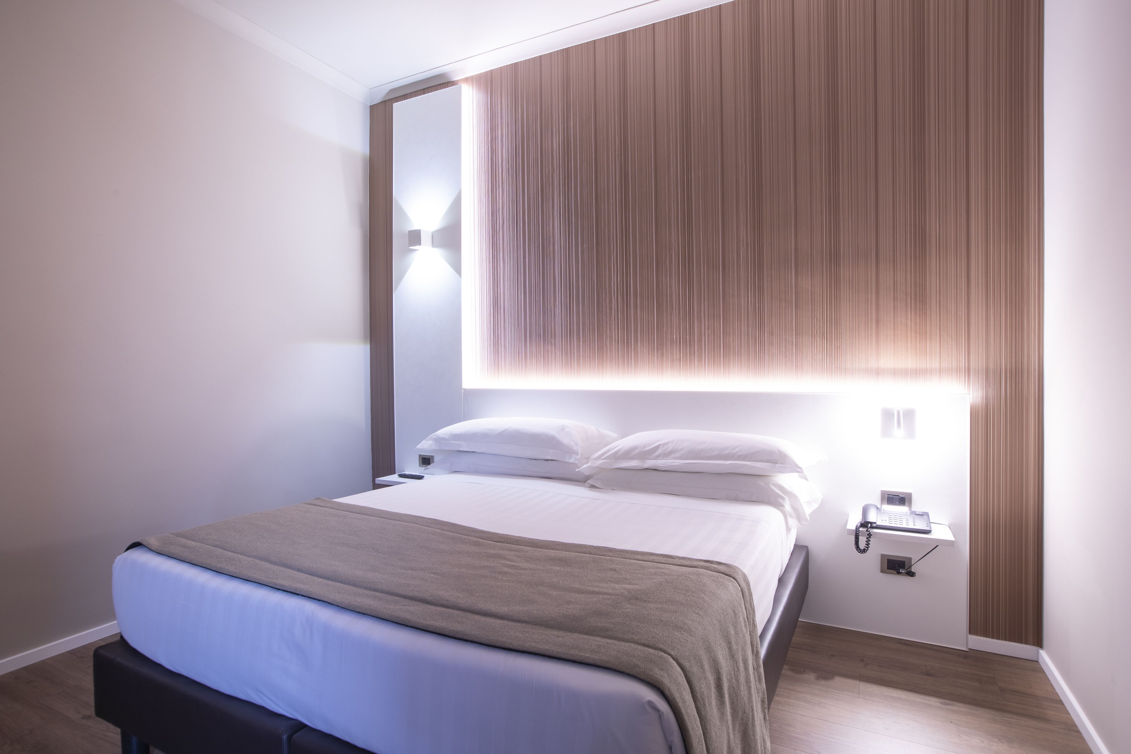 A modern hotel room with a neatly made bed, a bedside table with a phone, and a wall-mounted light fixture.