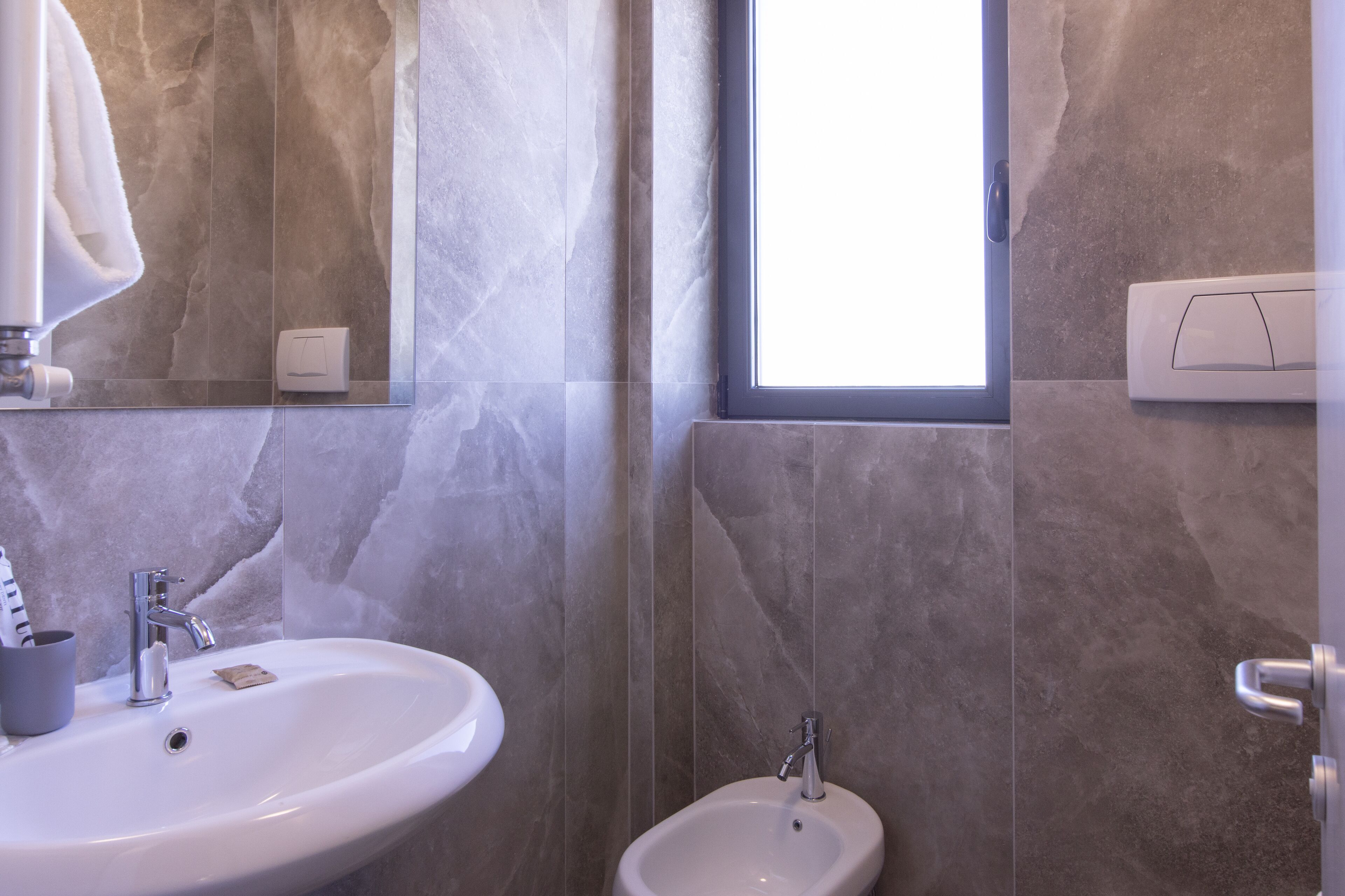 A bathroom with a large mirror, a sink, a bidet, and a window.