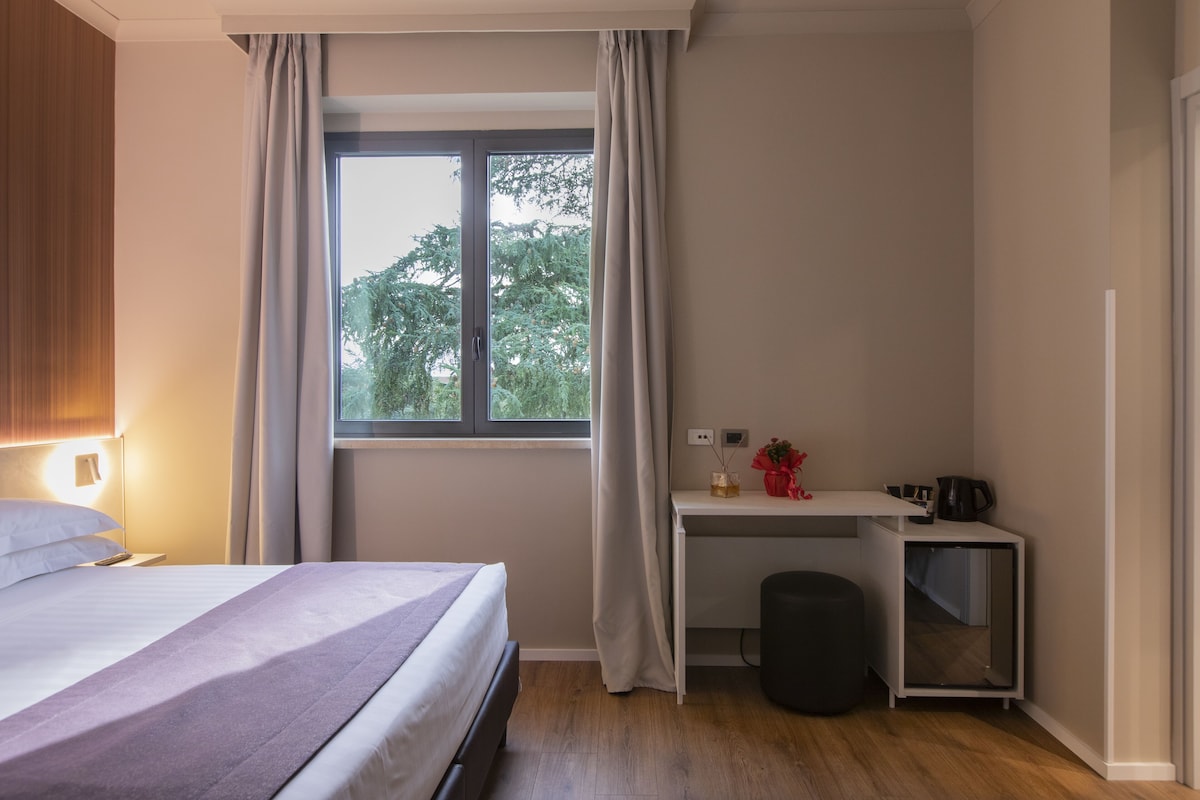 Una camera da letto con un letto, una scrivania, uno sgabello, una finestra con tende e vista sugli alberi.