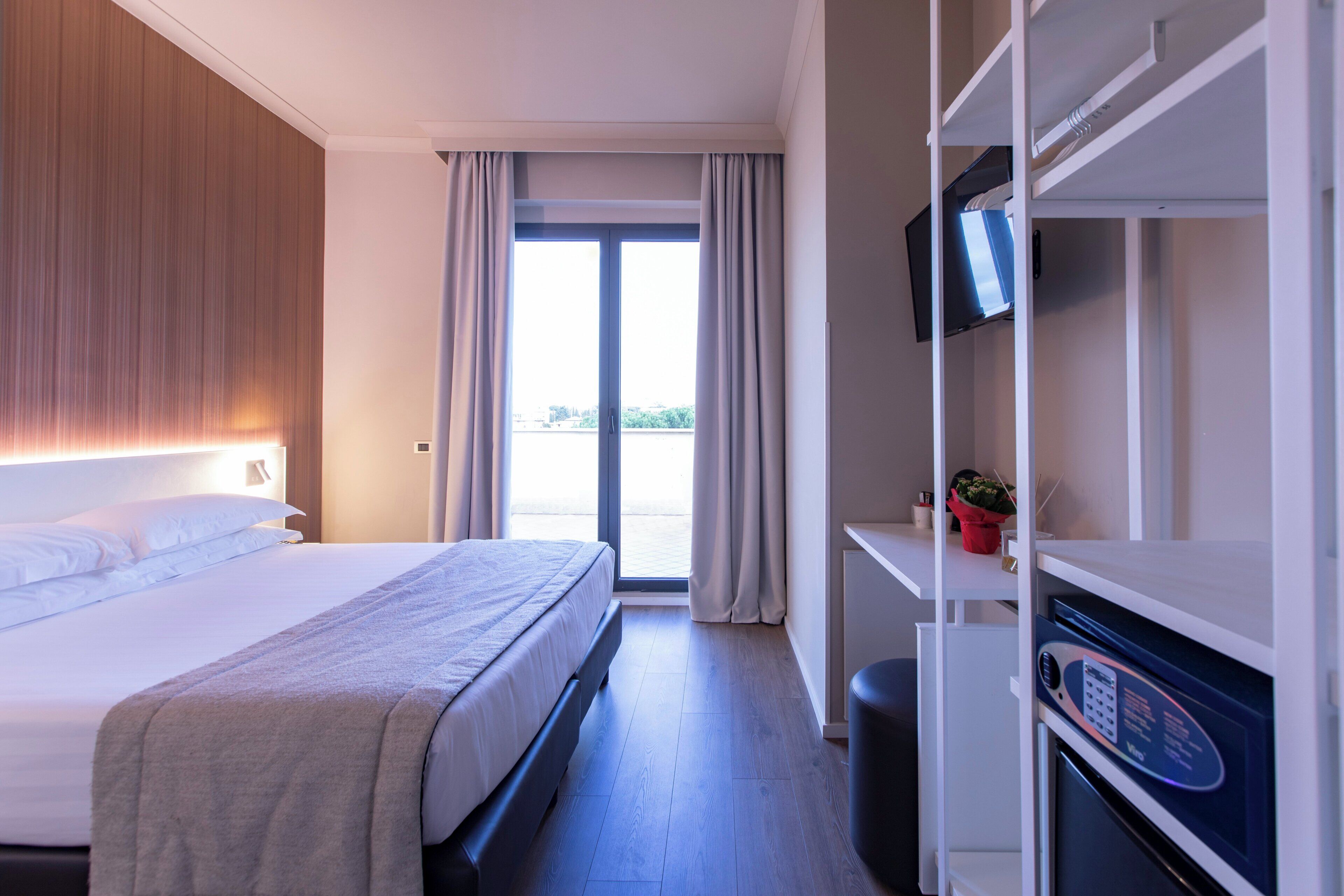 Une chambre d’hôtel avec un lit, une télévision, une petite table avec une plante et une vue sur l’extérieur grâce à une grande fenêtre.