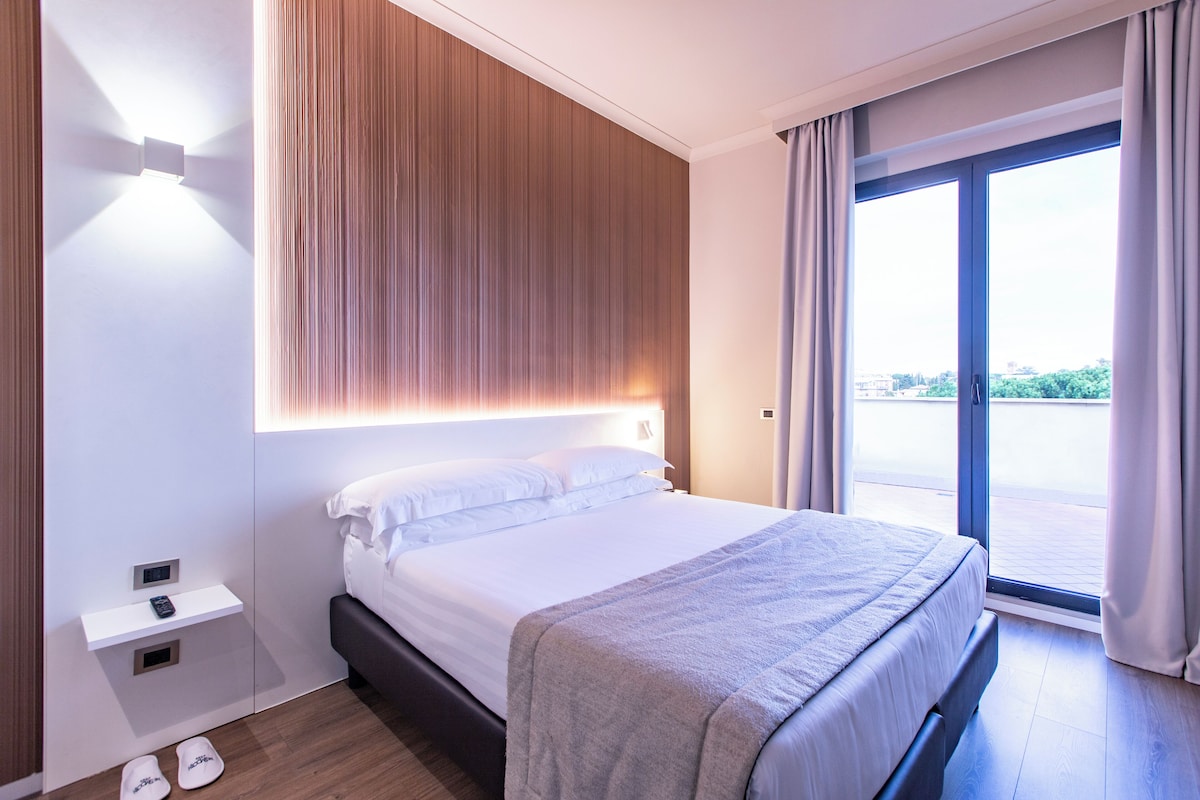 Camera d'hotel moderna con un letto grande, un comodino e vista sull'esterno tramite una porta scorrevole in vetro.