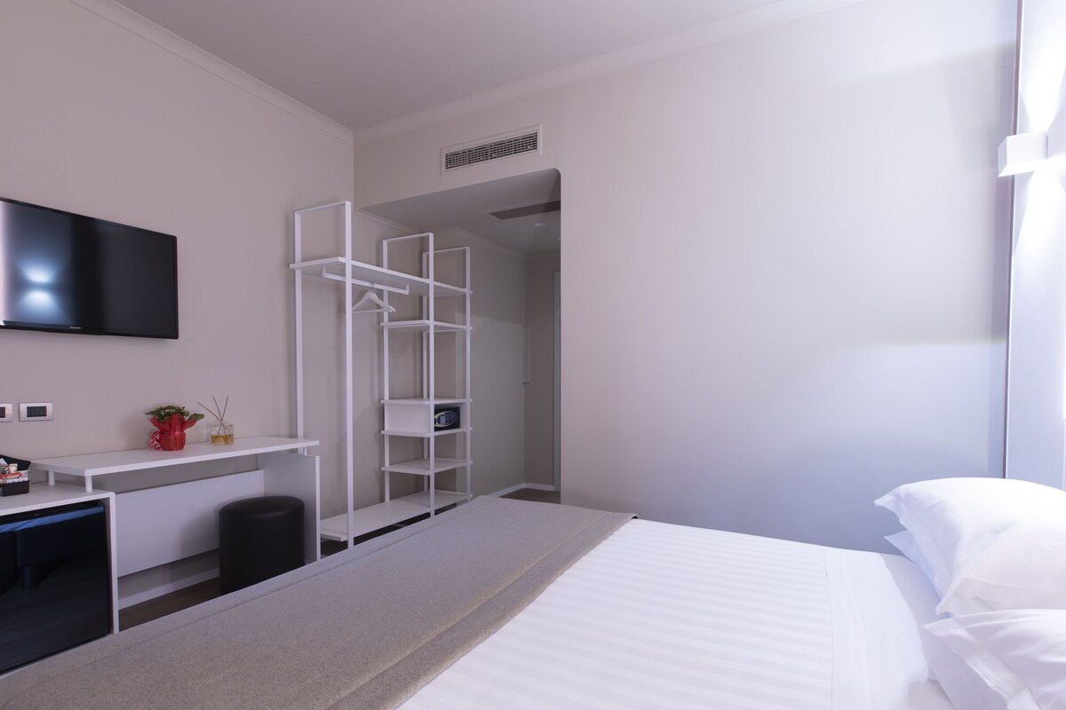 Una moderna camera d'hotel con un letto, una scrivania con televisore piatto, una scaffalatura bianca e un armadio a muro.