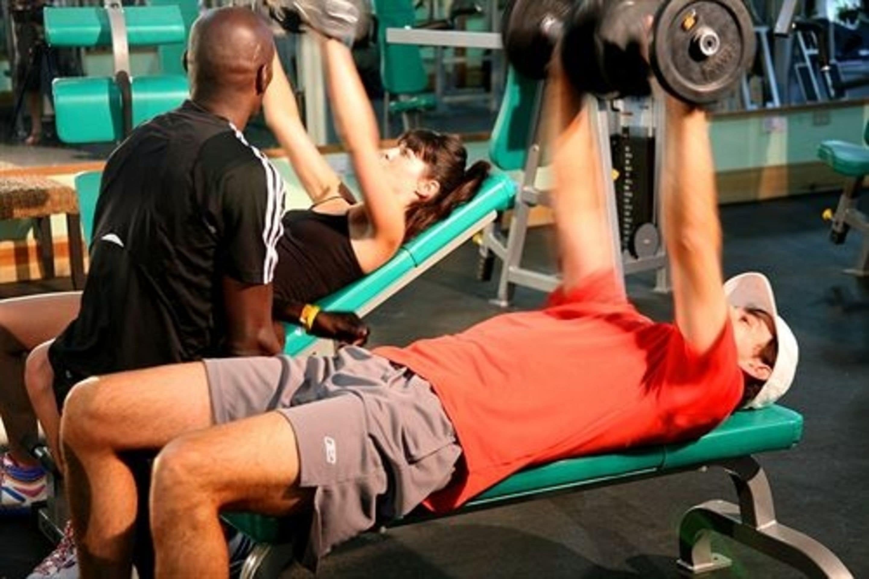 Una persona acostada en un banco dentro de un gimnasio, haciendo ejercicio con una máquina de pesas.