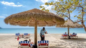 A beach with thatched umbrellas and striped beach chairs under a palm tree. - Sunset at the Palms Resort - Adults Only - All Inclusive (Negril)