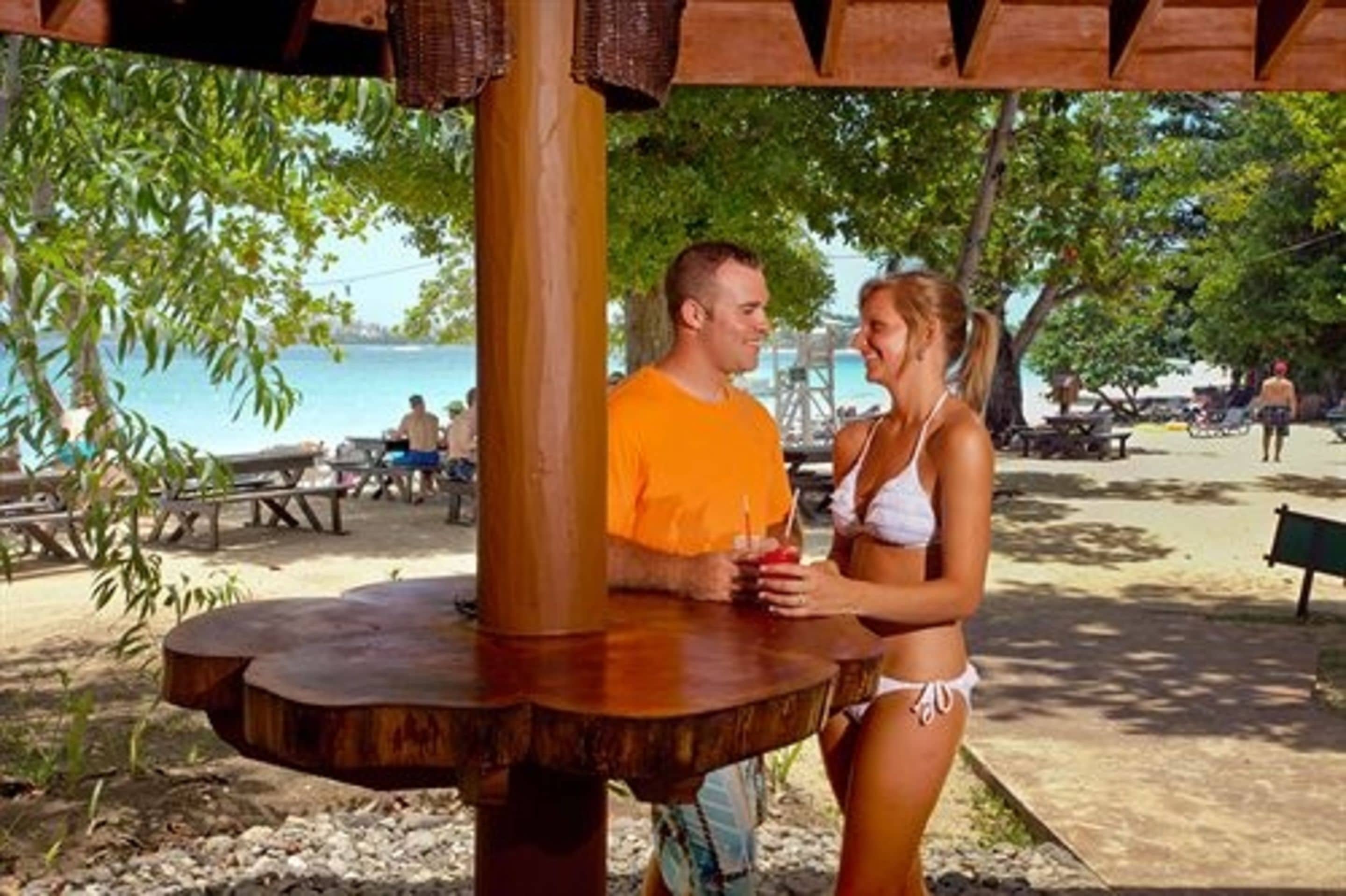 A wooden structure with a circular table, surrounded by a sandy area with trees and a body of water in the background.