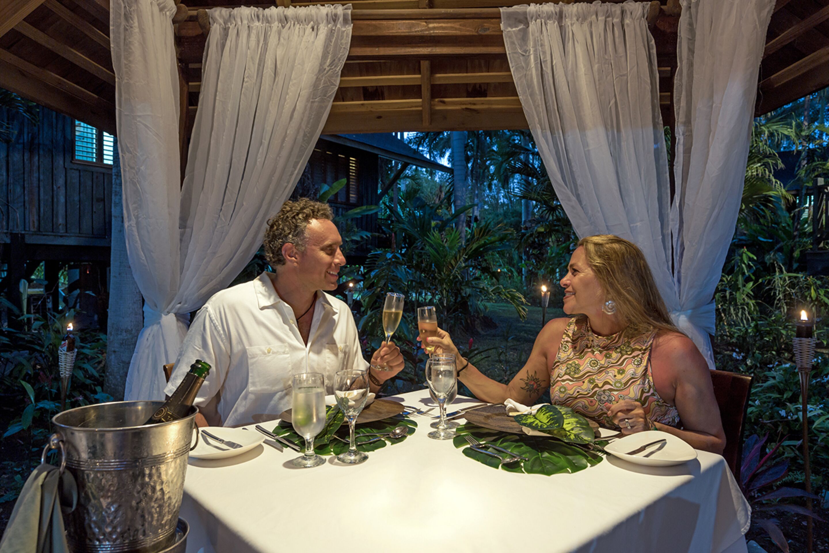 A couple toasting with champagne at an outdoor table with a view of tropical foliage.