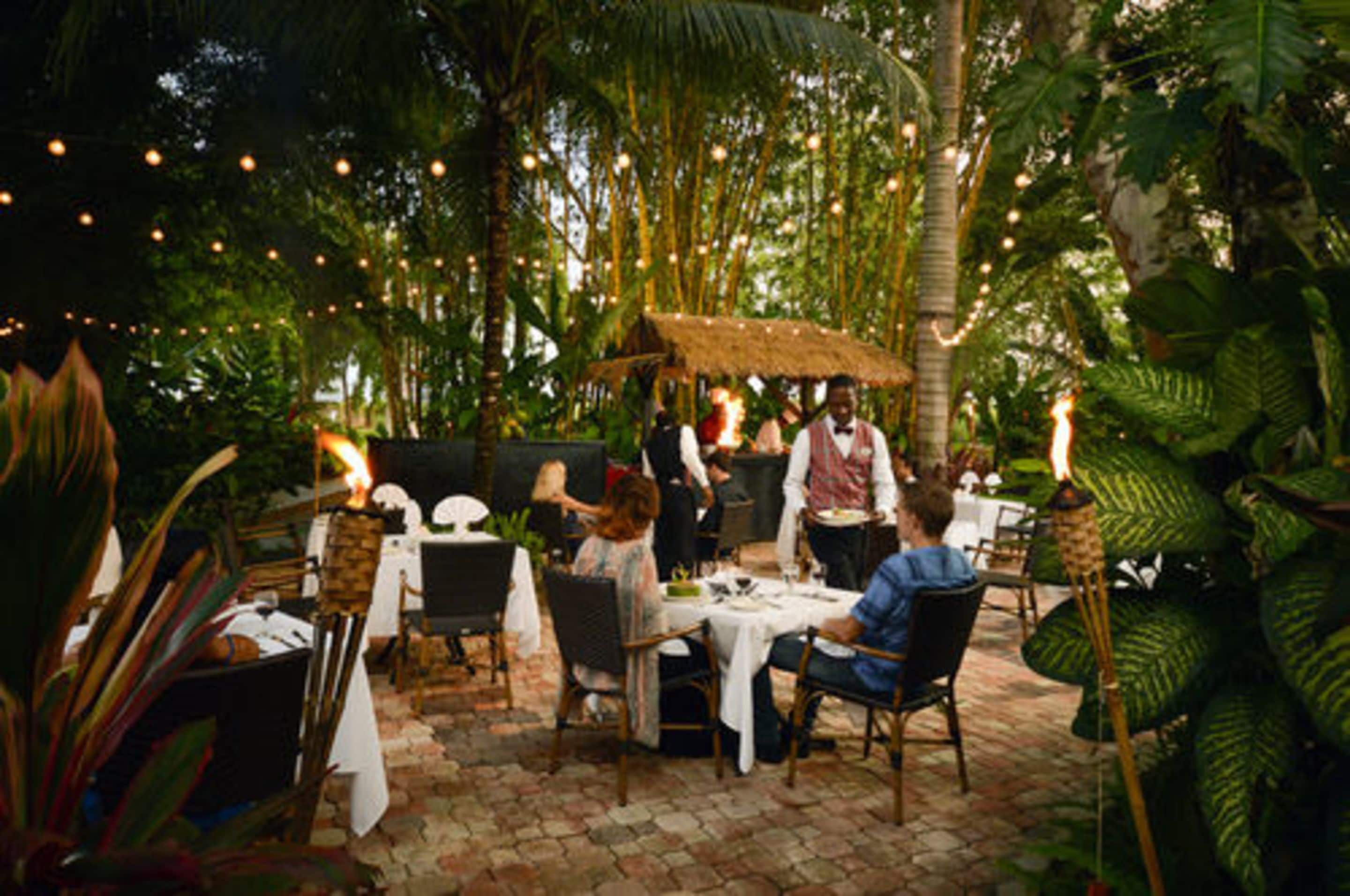 An outdoor dining area with tables and chairs, surrounded by lush greenery and string lights.