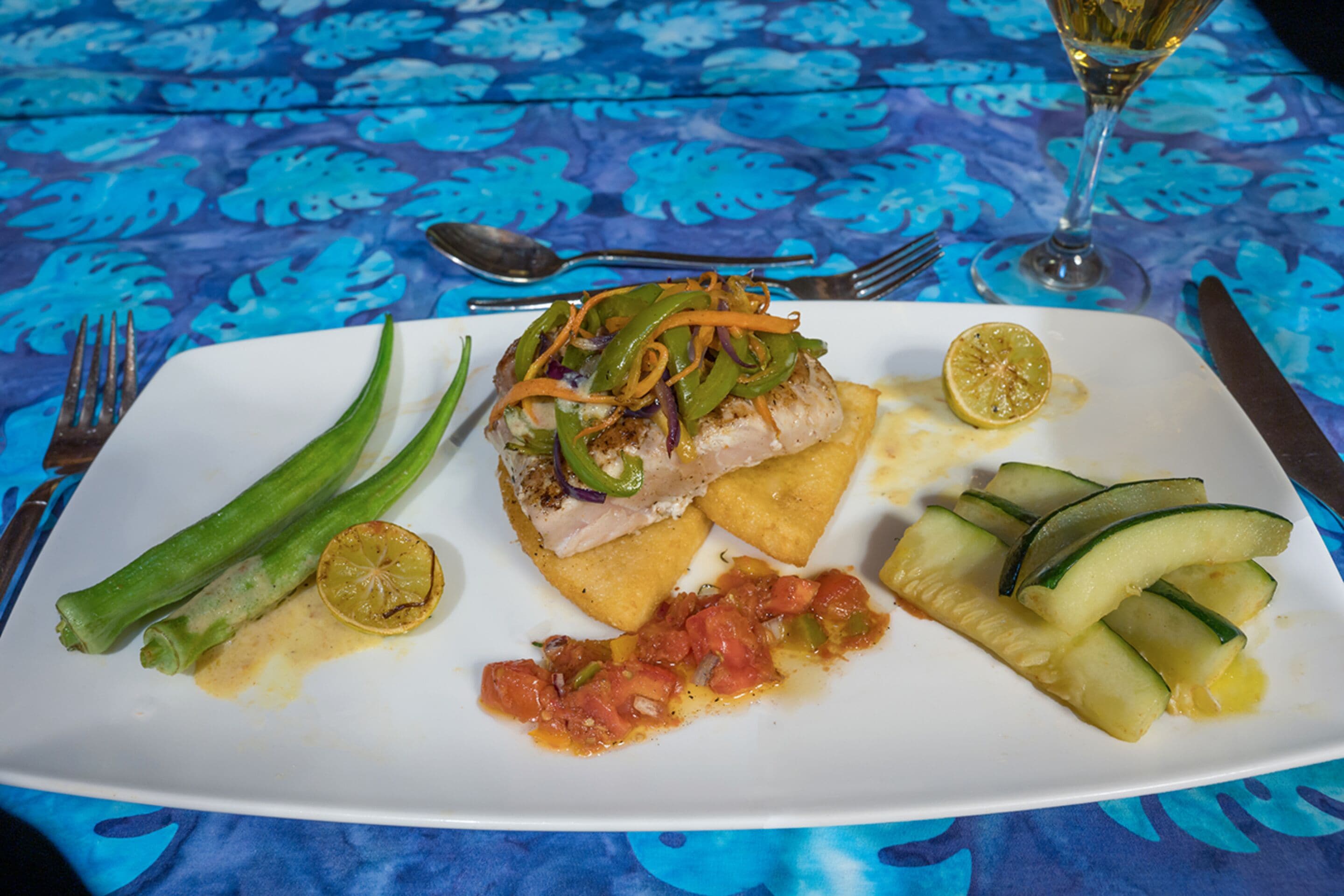 A white plate with a fish fillet, green vegetables, and a slice of lemon on a blue tablecloth.