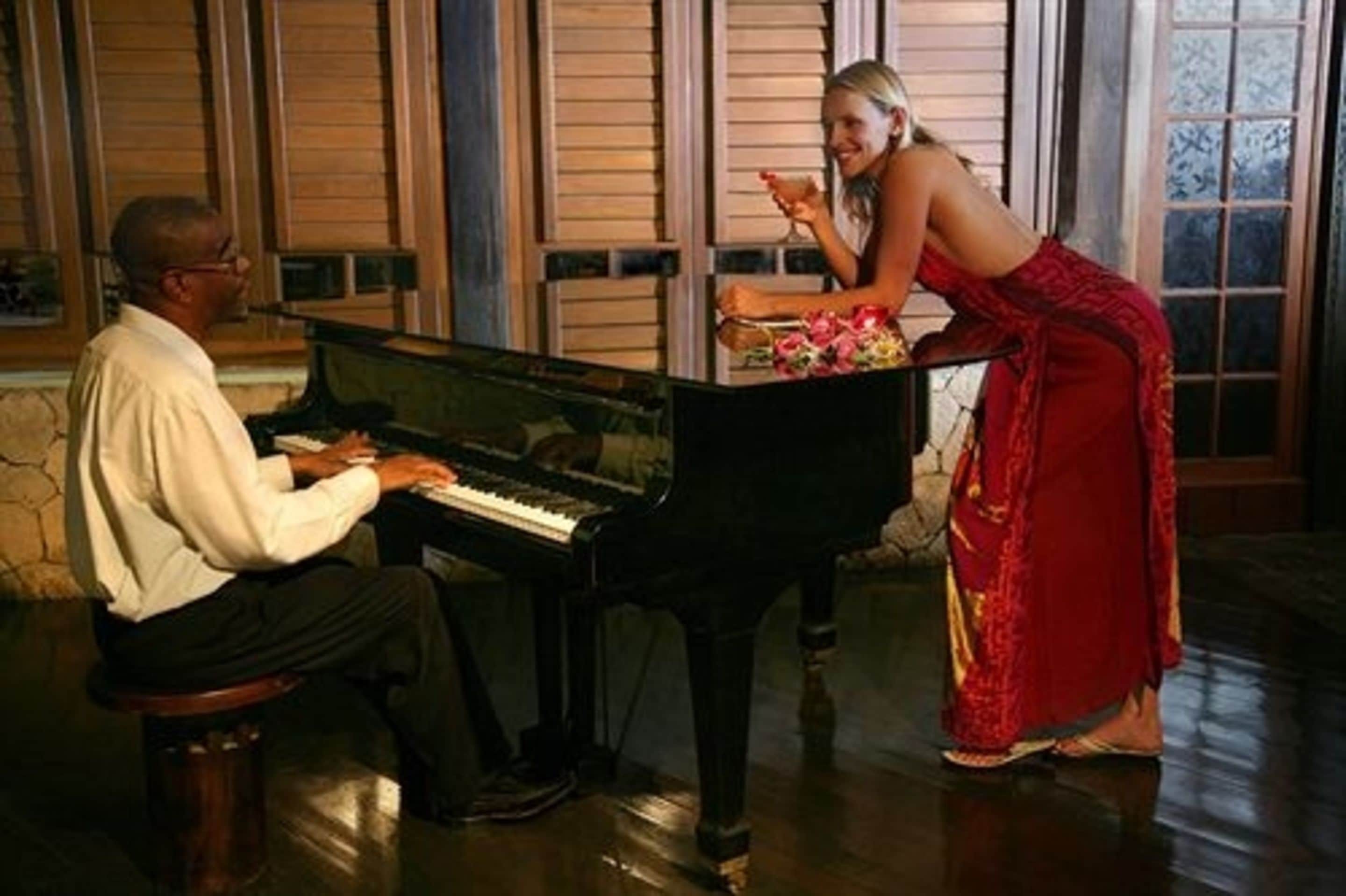 Un hombre tocando el piano en una habitación, mientras una mujer está cerca sosteniendo una bebida.
