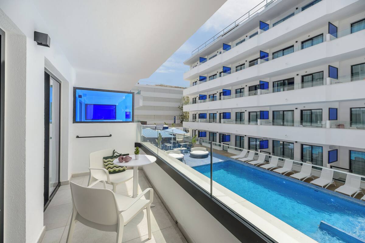 Un balcón moderno de hotel con piscina, mobiliario blanco y vistas al interior del edificio.