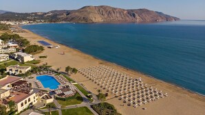 Vista aérea de un resort costero con una gran piscina, sombrillas de playa y una montaña al fondo.