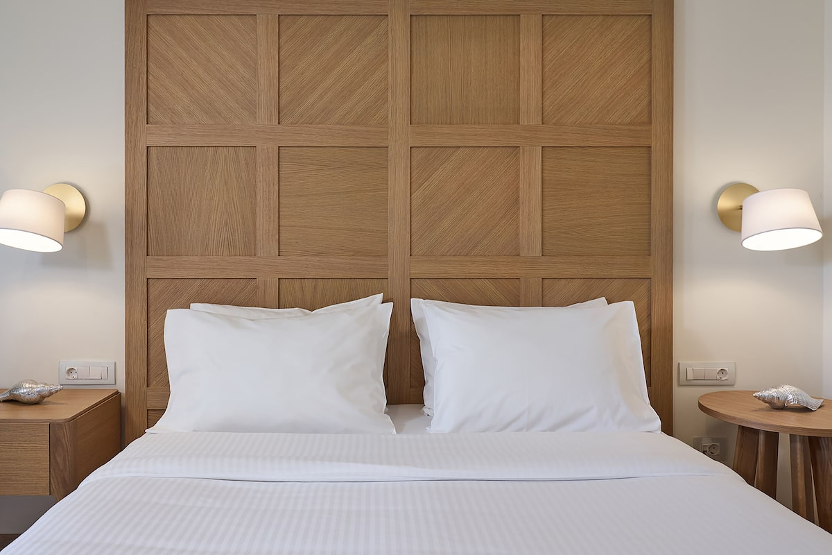 Una habitación de hotel con un gran cabecero de madera, dos almohadas blancas, una cama bien hecha, dos mesitas de noche con lámparas y una pequeña mesa redonda con una lámpara.