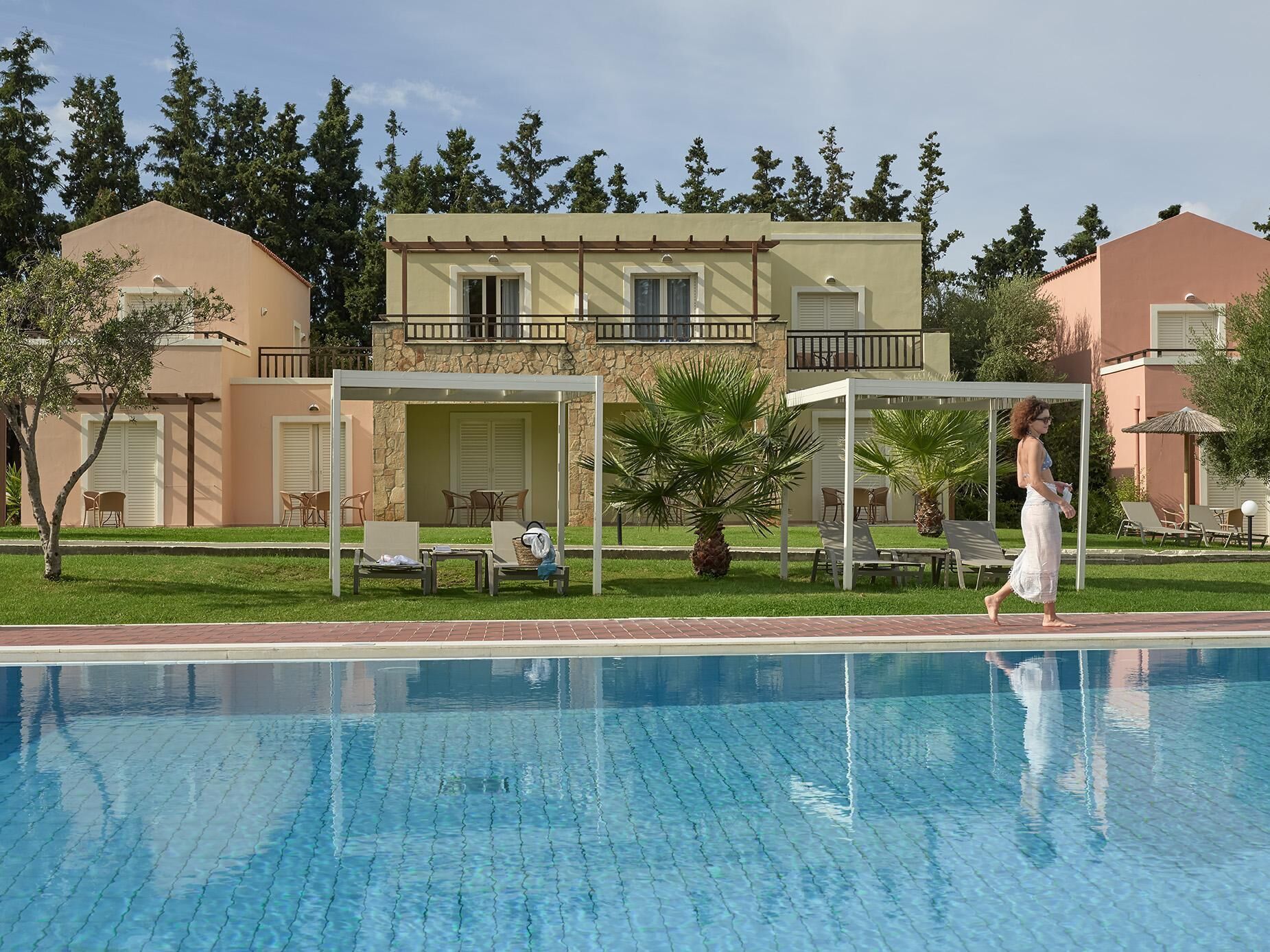 A swimming pool with lounge chairs and umbrellas, a person walking, and a building with balconies and outdoor seating.