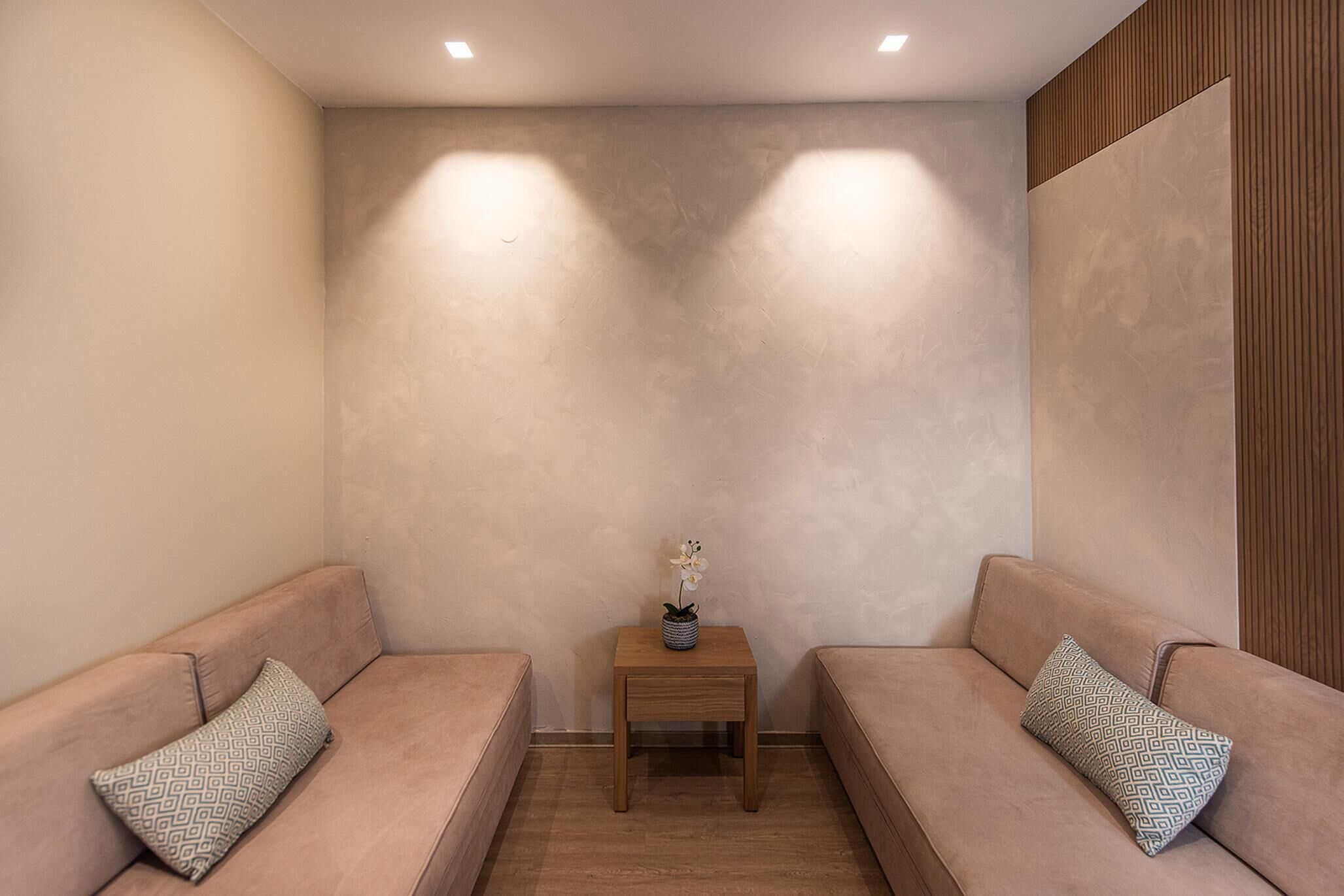A room with two beige sofas, a small wooden side table with a vase of flowers, and a textured wall.
