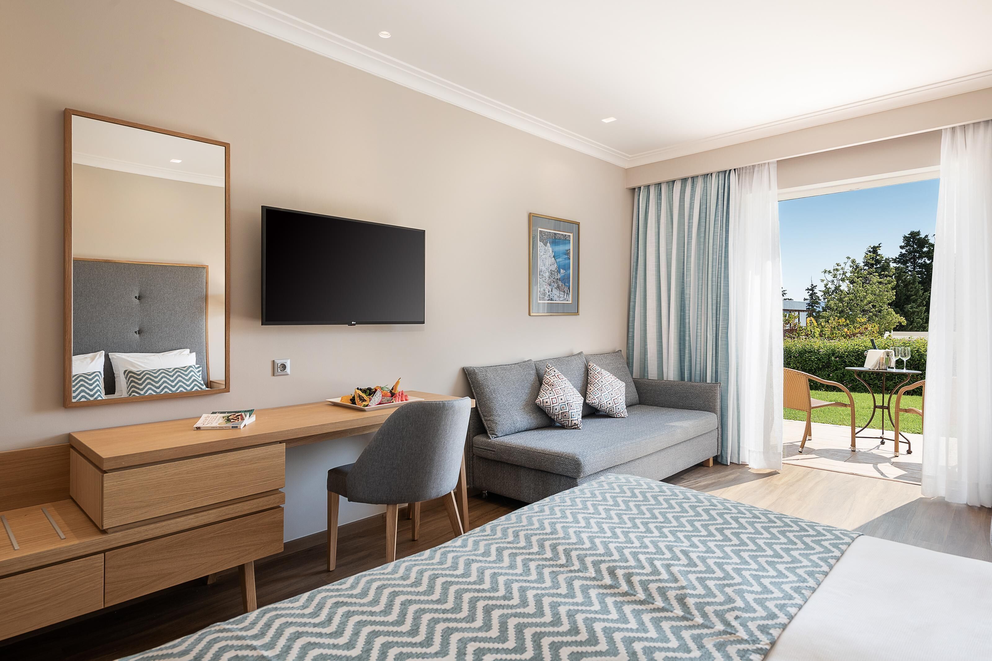 A modern hotel room with a bed, a desk with a chair, a sofa, a TV, and a balcony with outdoor seating.