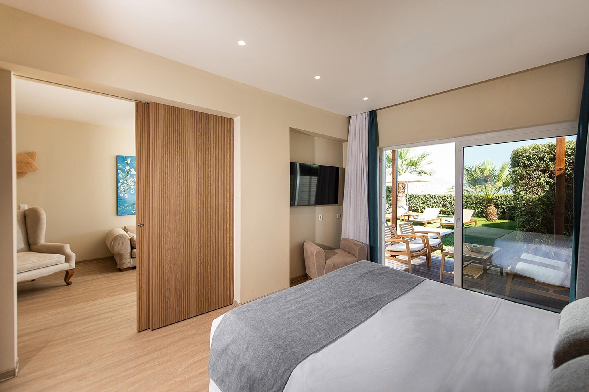 Una habitación de hotel moderna con una cama grande, un televisor y un ventanal corredera que da a un balcón con mobiliario exterior.