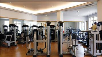 A gym with various exercise machines, a man using a treadmill, and large windows allowing natural light.