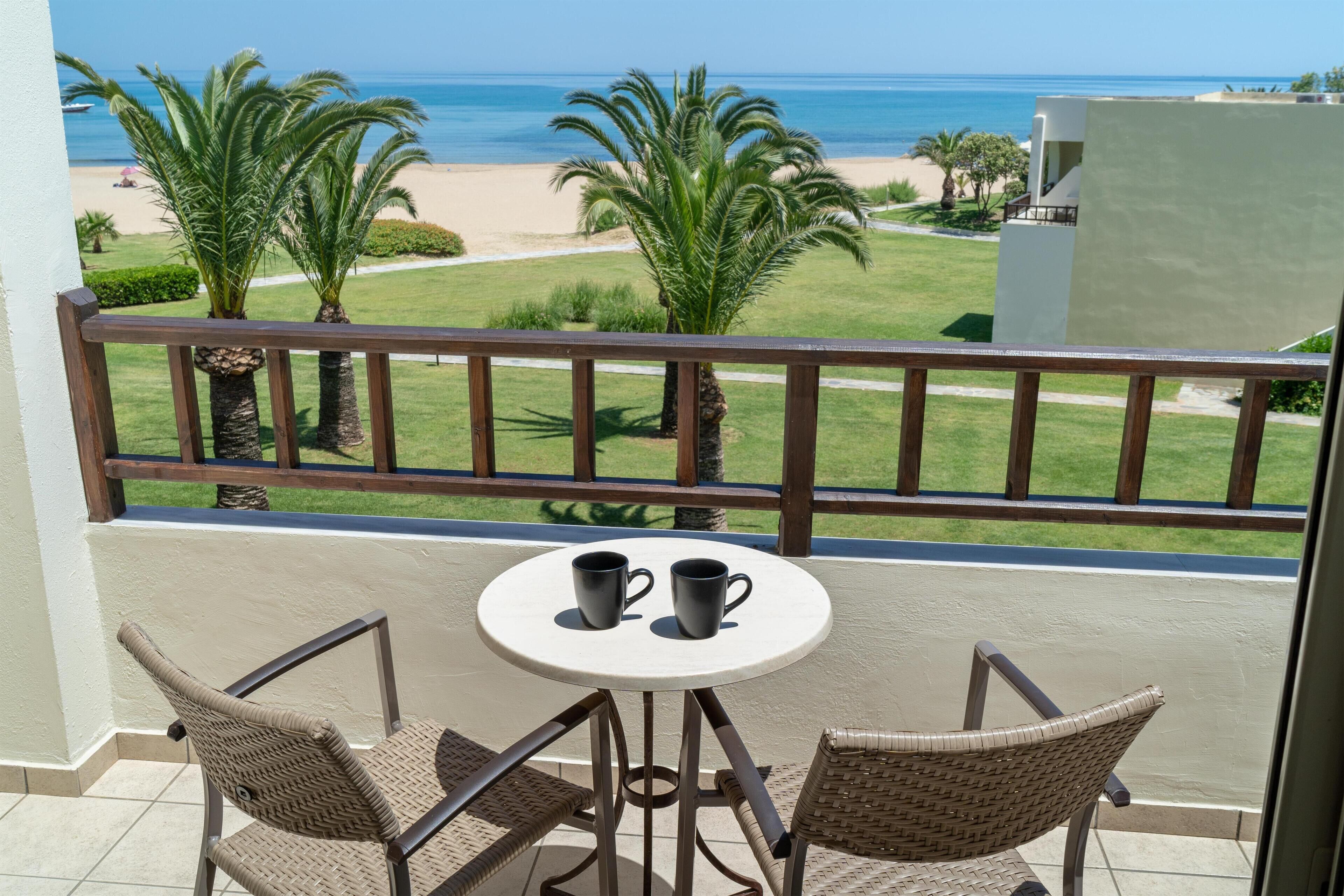 Un balcon avec deux fauteuils en osier, une petite table ronde et deux tasses noires. La vue comprend une plage et des palmiers.