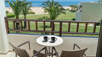 Ein Balkon mit zwei Korbstühlen, einem kleinen runden Tisch und zwei schwarzen Tassen. Blick auf Strand und Palmen.