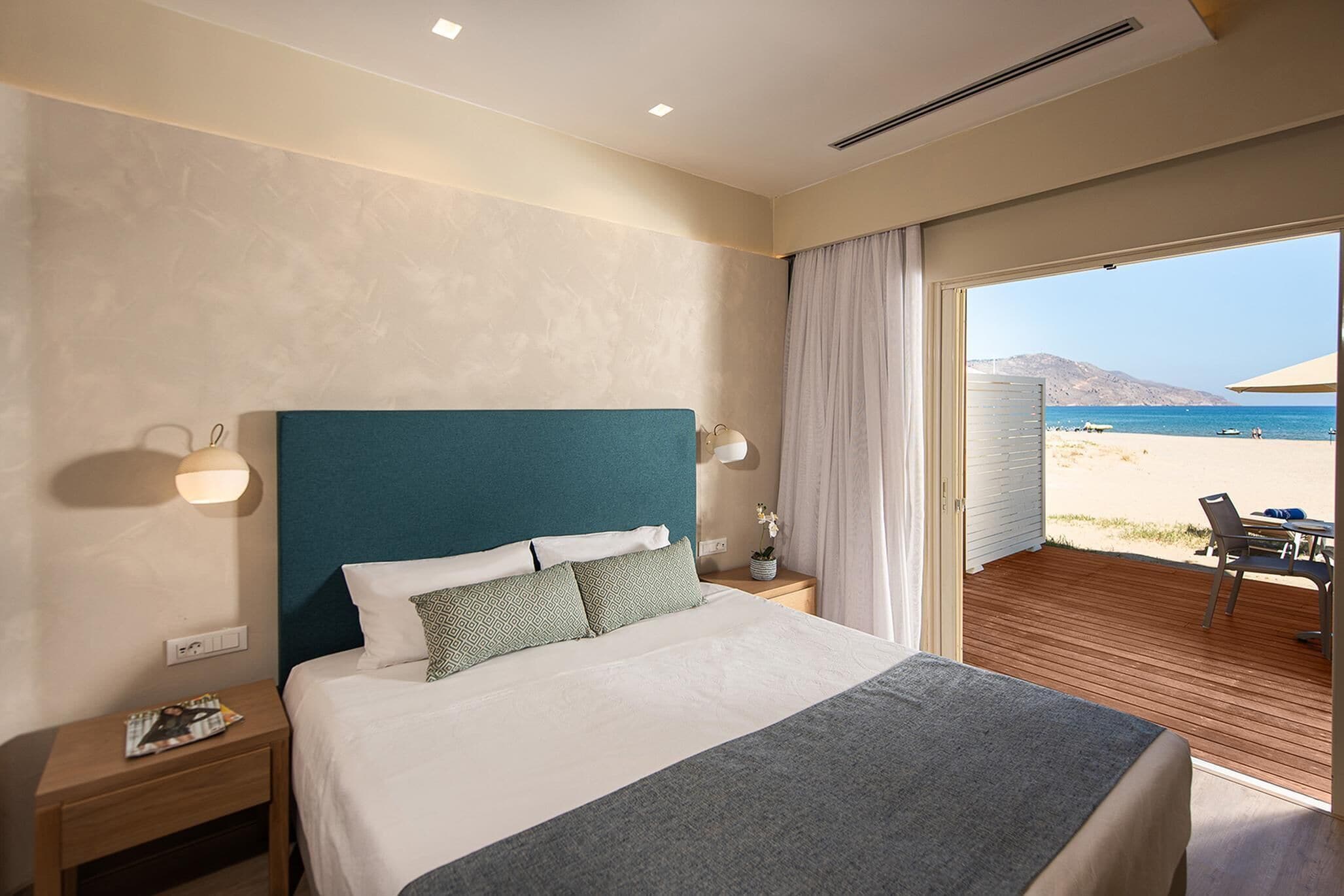 A modern hotel room with a large bed, a wooden floor, a view of the beach, and a balcony with outdoor seating.